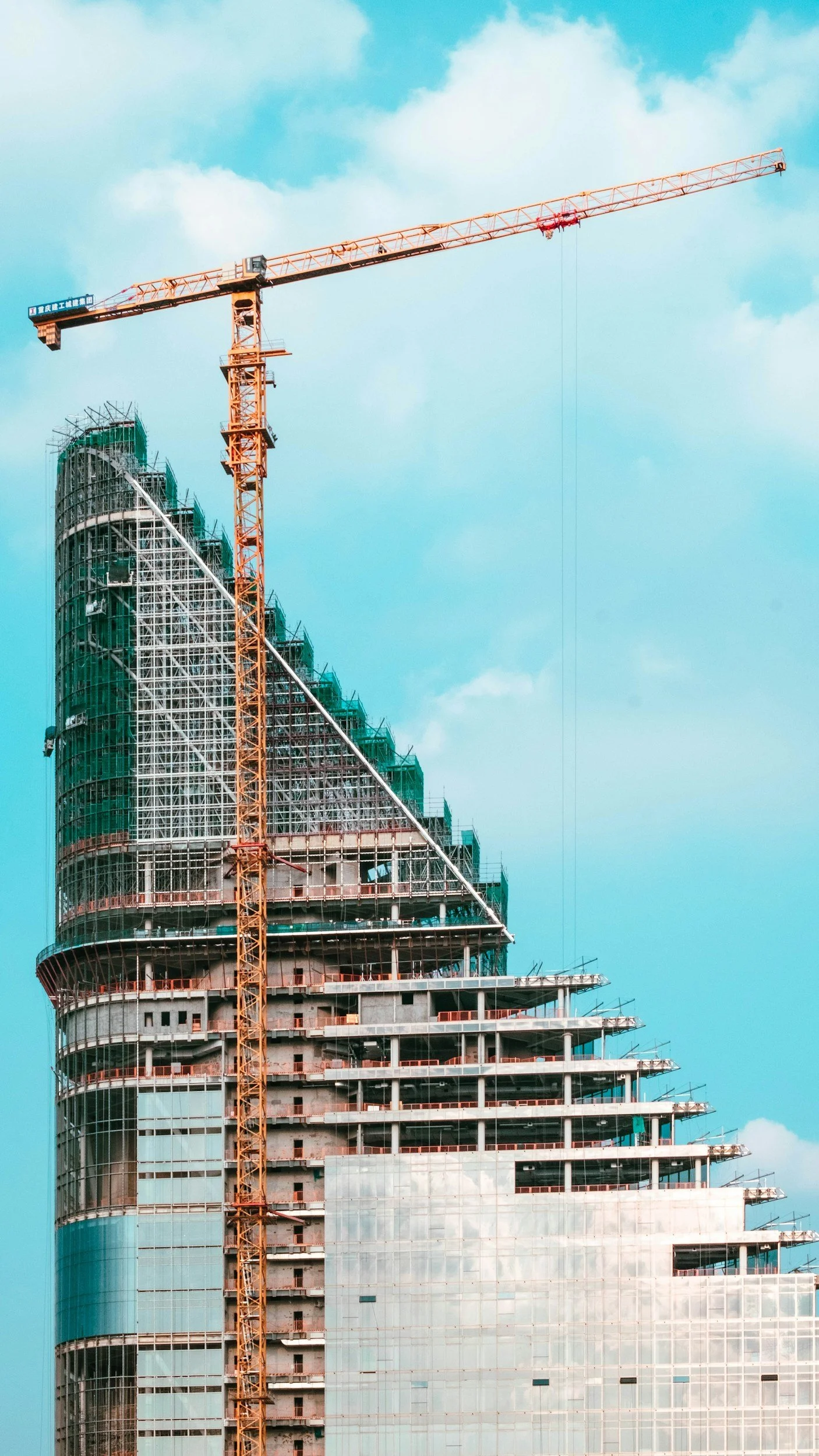 A high-rise building under construction with scaffolding and a tower crane against a blue sky with clouds.