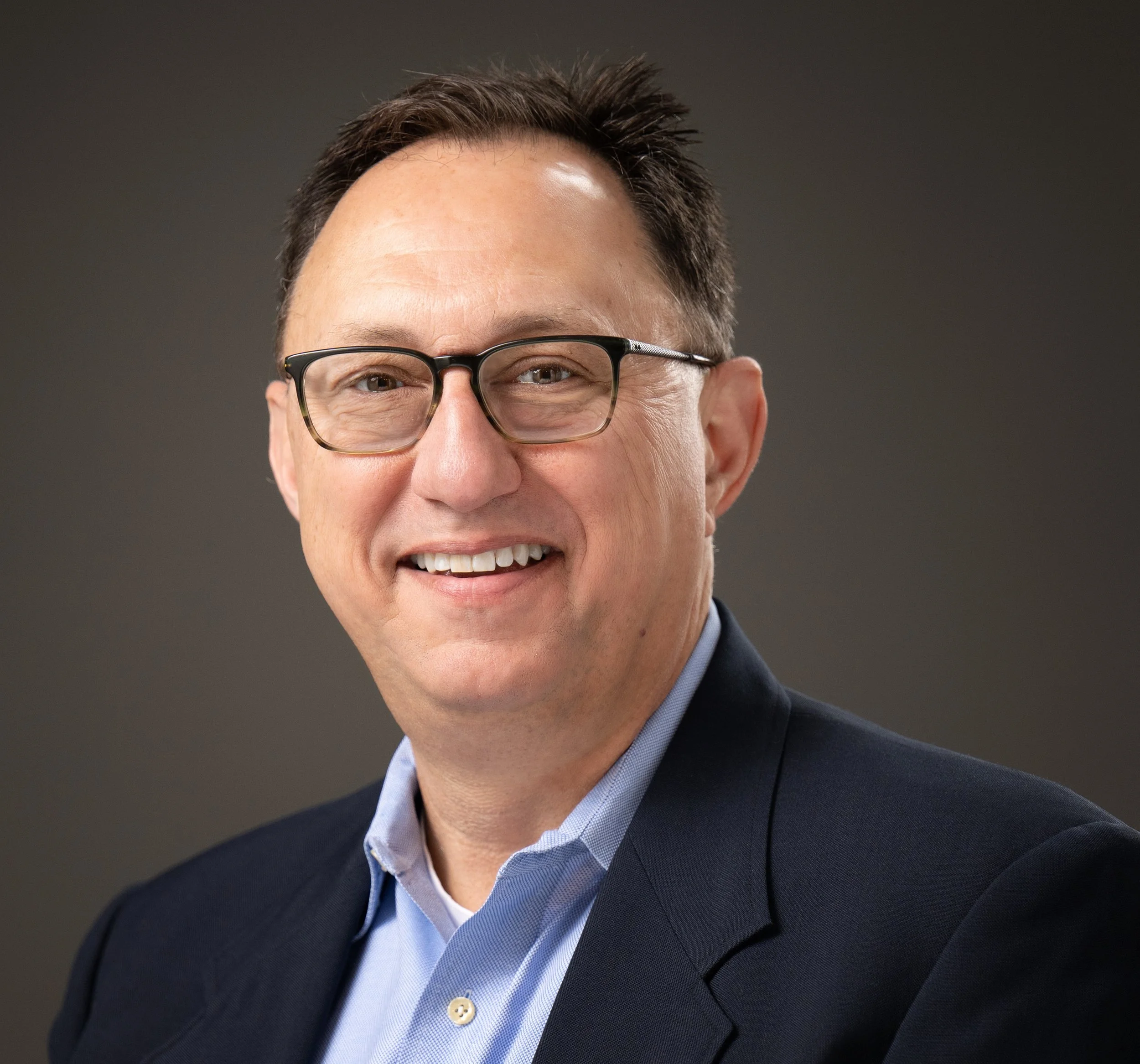 A man with glasses, dark hair, wearing a navy blazer and light blue shirt, smiling against a neutral background.