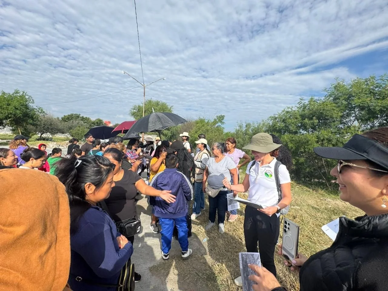 Grupo de personas en un campo abiertos, algunas con sombreros y paraguas, participando en una actividad de senderismo o campo, con cielo parcialmente nublado y árboles en el fondo.