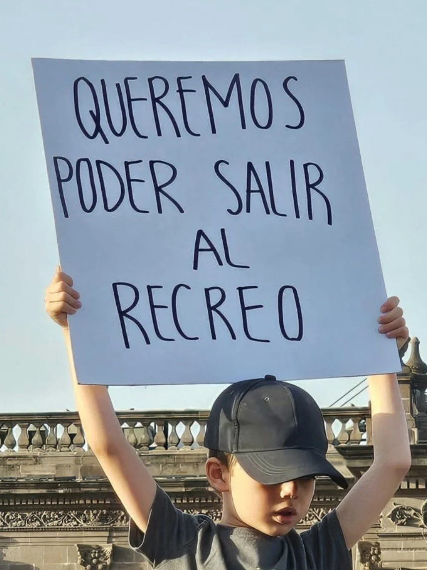 Niño sosteniendo cartel que dice "Queremos poder salir al recreo" en una protesta al aire libre.