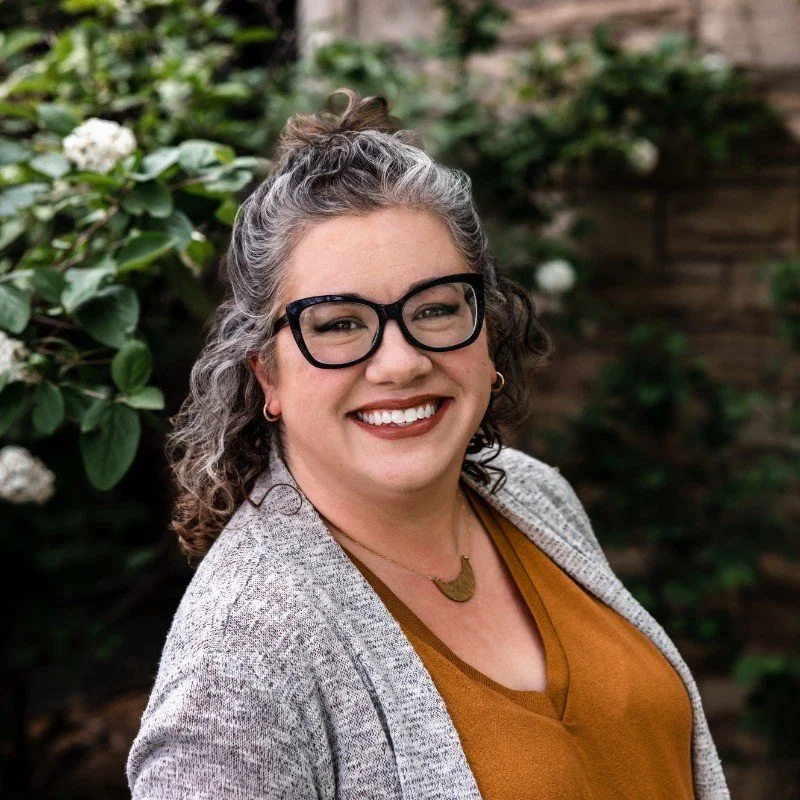 A woman with curly gray hair, wearing black glasses, a gold necklace, a brown top, and a gray cardigan, smiling outdoors with green plants and white flowers in the background.