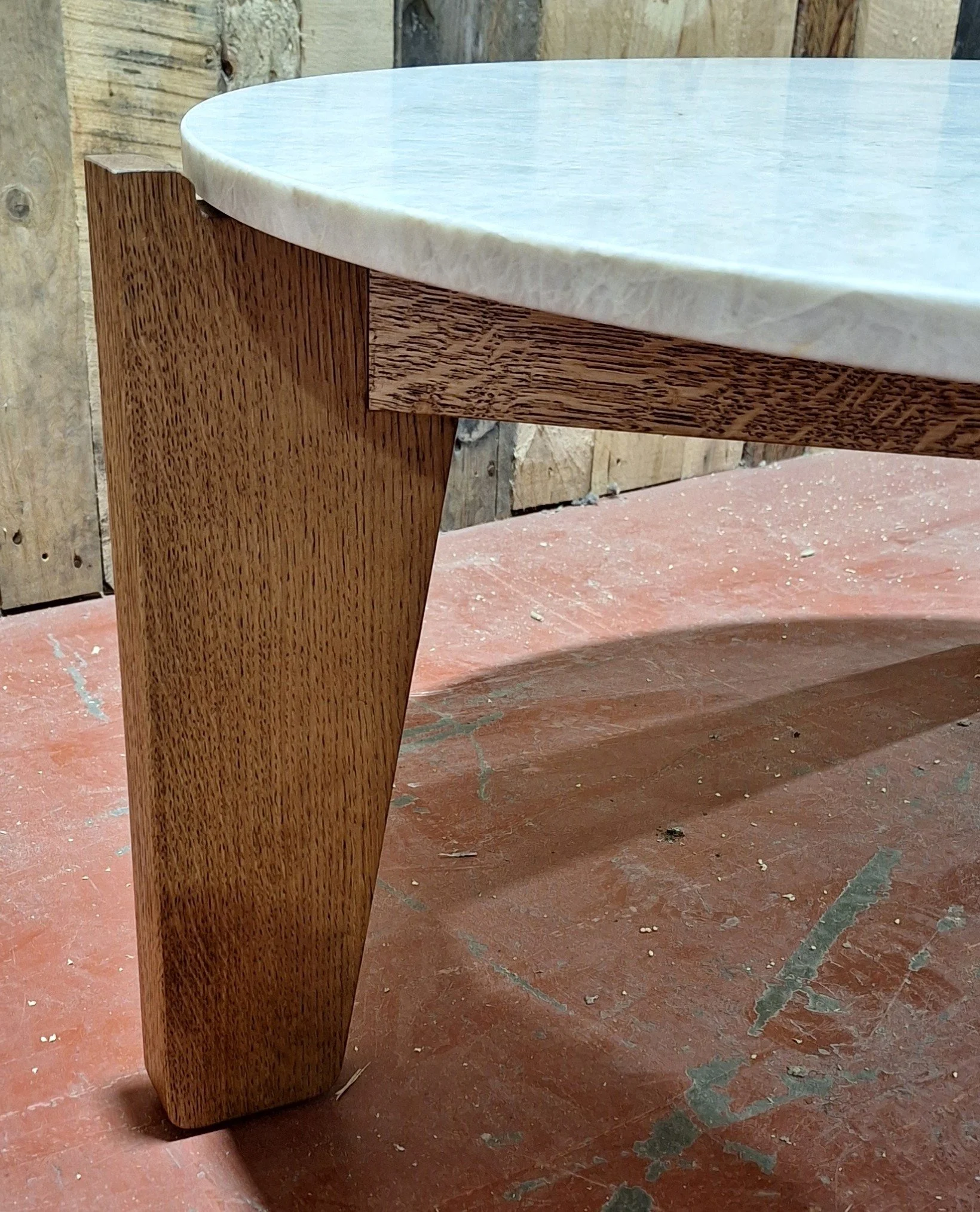 Close-up of a wooden table leg and edge, with a light-colored stone or marble top, placed on a dusty reddish-brown floor with visible scratches and dust, and a wooden wall in the background.
