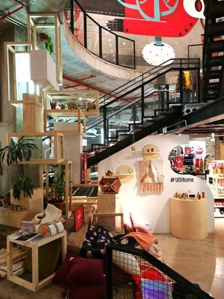 Interior of a modern, multi-level store with wooden shelves, plants, a staircase, and various decorative items.