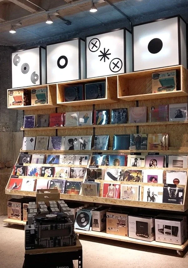 Vinyl records displayed on wooden shelves in a store, with colorful album covers and some boxed turntables on the lower shelf, under three large illuminated graphic panels.
