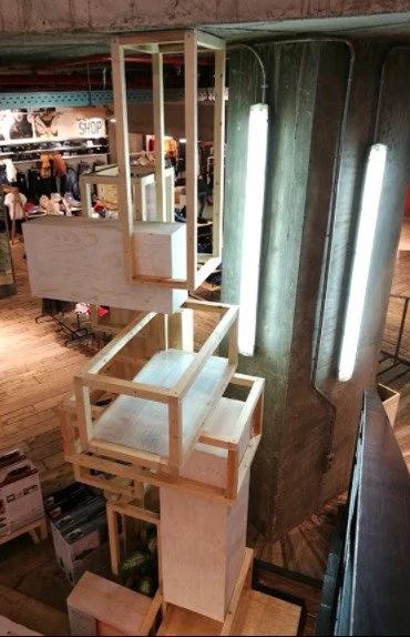 A tiered wooden display stand with multiple open square frames, placed next to a large, curved metallic wall with vertical lighting fixtures, in a retail store.