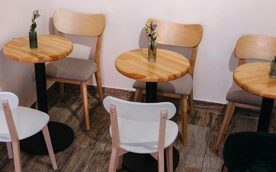 Three round wooden tables with small vases holding flowers, surrounded by a mix of wooden and white chairs in a cafe or restaurant setting.