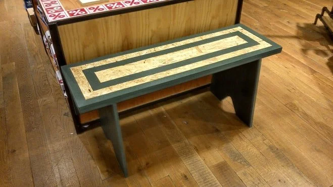 A rectangular wooden bench with a painted geometric pattern on top, situated on wooden flooring inside a store.