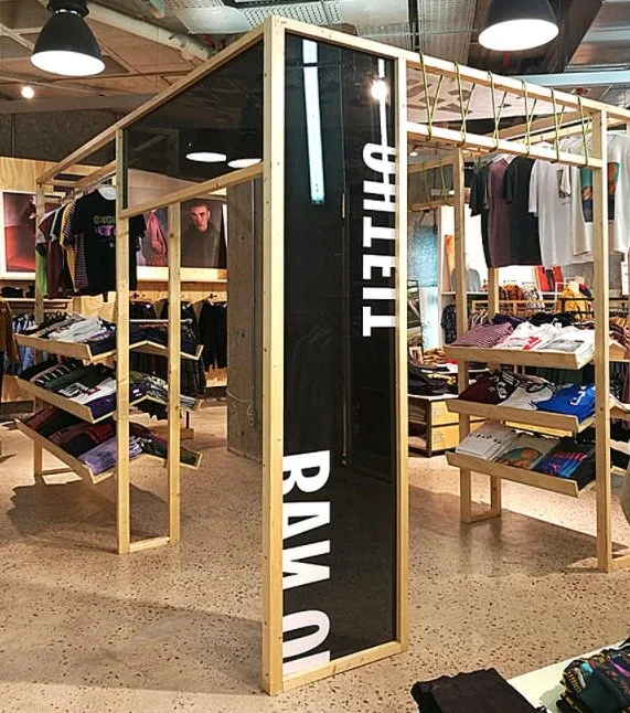 A clothing store display made of light wooden frames with hanging clothes and neatly folded folded shirts and pants on shelves. The store has industrial ceiling and lighting. A vertical mirror with white letters is mounted on the display.