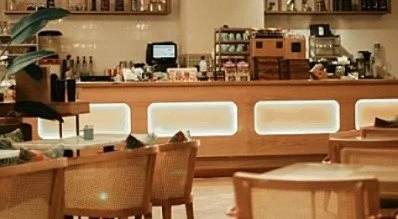 A cozy coffee shop interior with a wooden counter, illuminated panels, and seating area with wicker chairs.