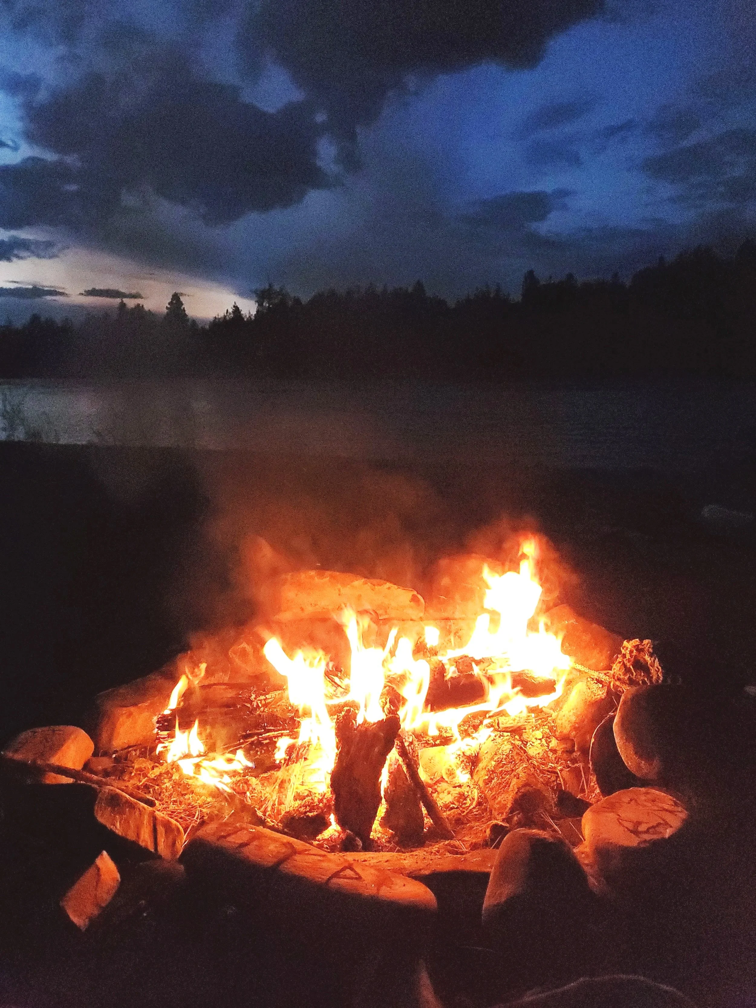 A campfire surrounded by rocks near a body of water during dusk or night, with dark clouds and trees in the background.