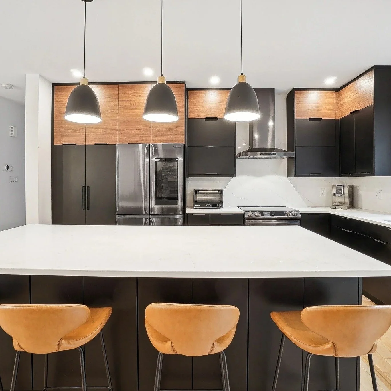 Cuisine moderne intemporelle avec un îlot central blanc, des plans de travail blancs, des armoires noires et en bois, des appareils électroménagers en acier inoxydable, trois suspensions noires au plafond et trois chaises en bois autour de l'îlot.