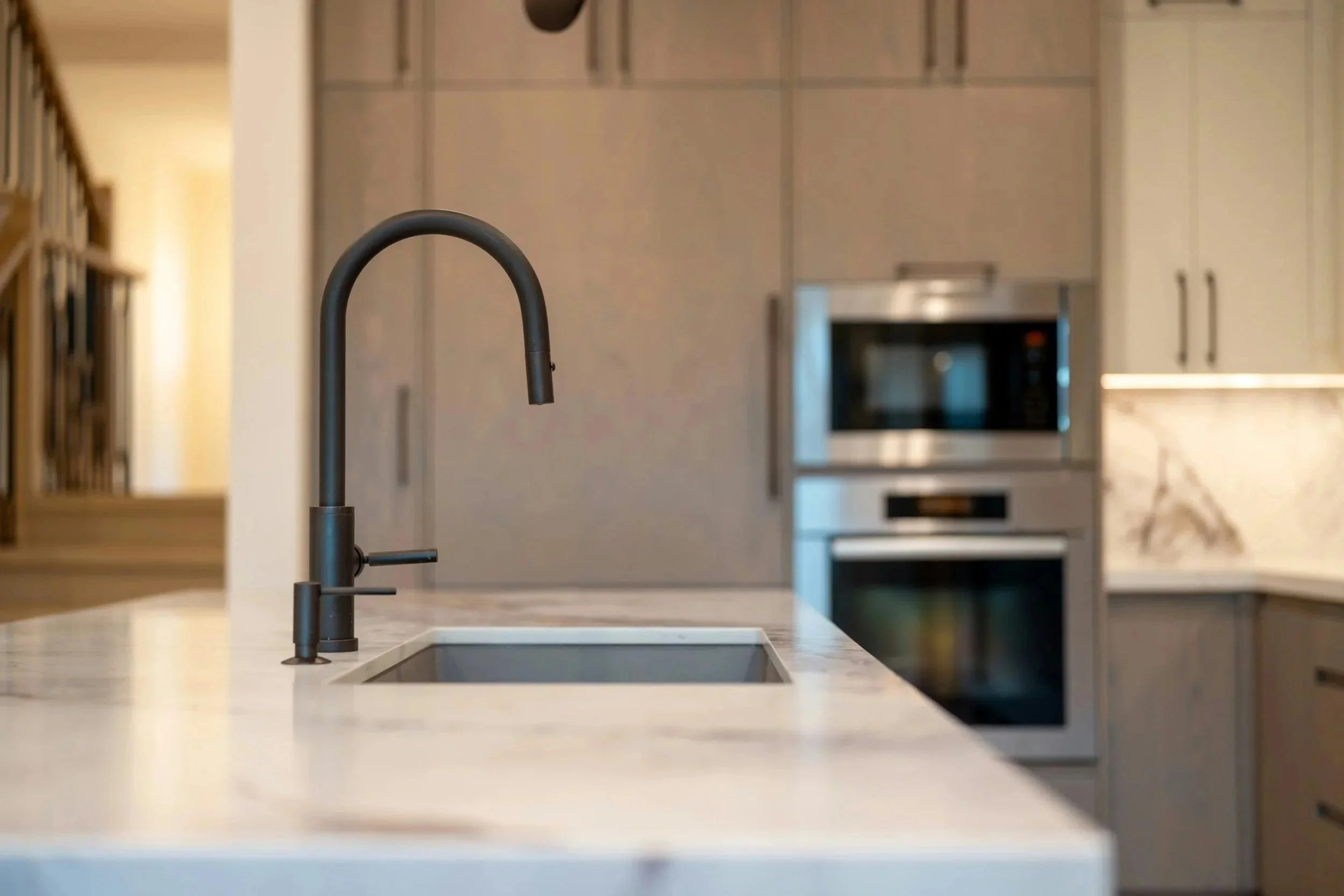 Vue d'une cuisine moderne avec un évier noir et un robinet noir placé sur un plan de travail en marbre blanc, avec des appareils électroménagers intégrés en arrière-plan.