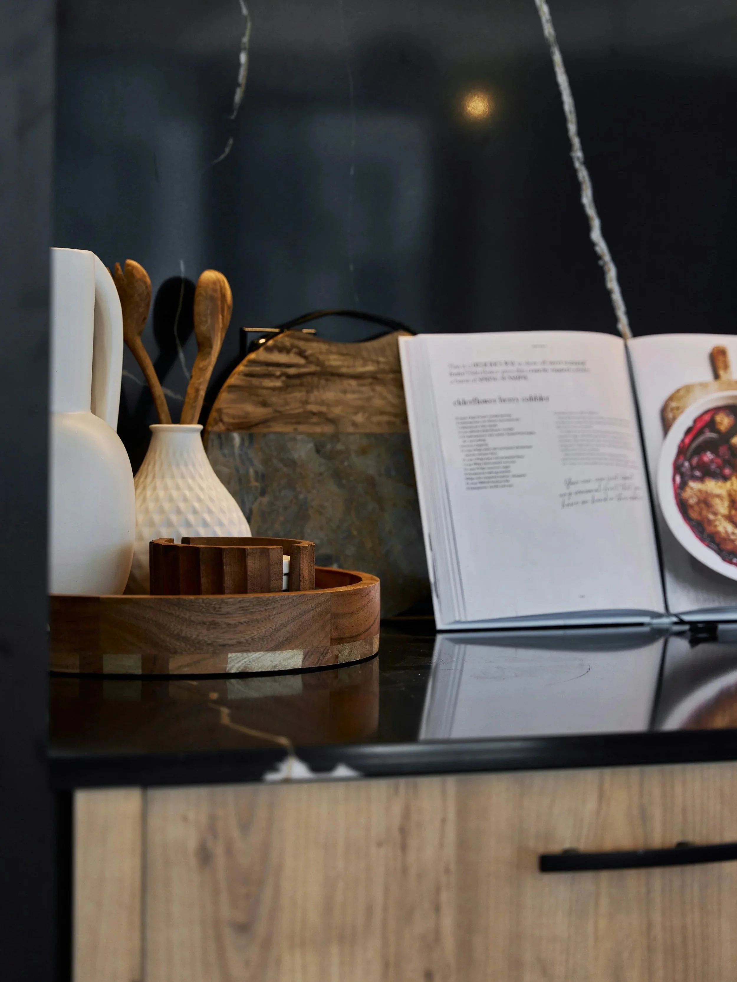 Un tableau de cuisine avec un vase blanc contenant des cuillères en bois, un plateau en bois, un livre de recettes ouvert, et une planche en pierre avec un manche en bois, contre un mur noir en quartz riche.