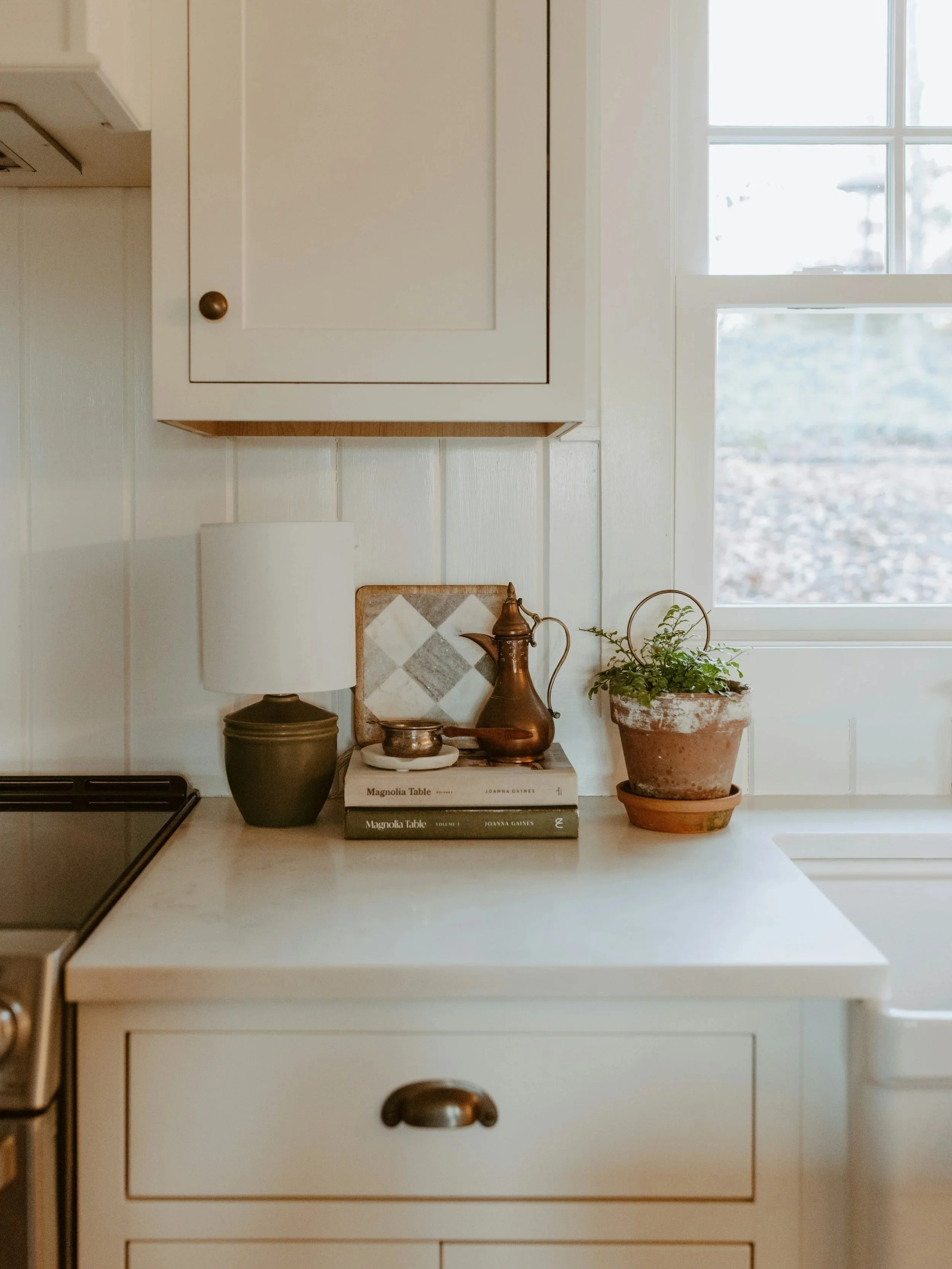 Coin de cuisine avec une étagère blanche, une lampe à base noire, un livre intitulé "Magnolia Table", un vase en cuivre, une petite assiette, un pot de terre avec une plante, une fenêtre laissant entrer la lumière naturelle.