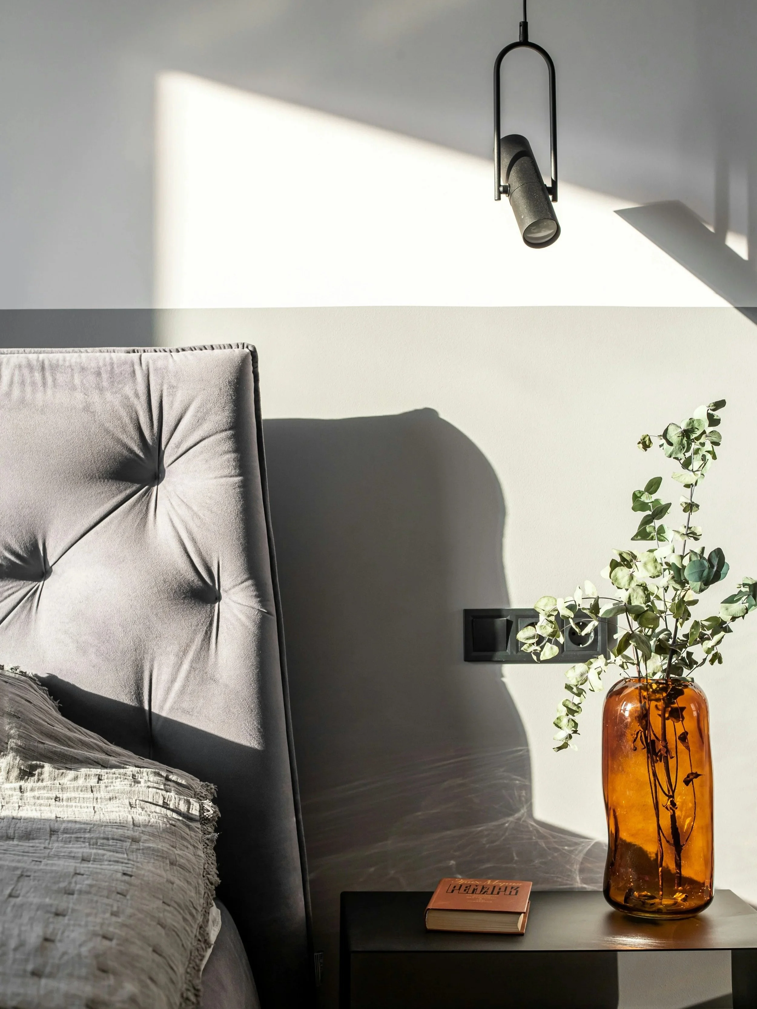 Une tête de lit en tissu gris clair, une lampe suspendue noire, une table de nuit noire avec un livre et un vase en verre ambre avec des feuilles vertes, dans une pièce lumineuse et confortable.