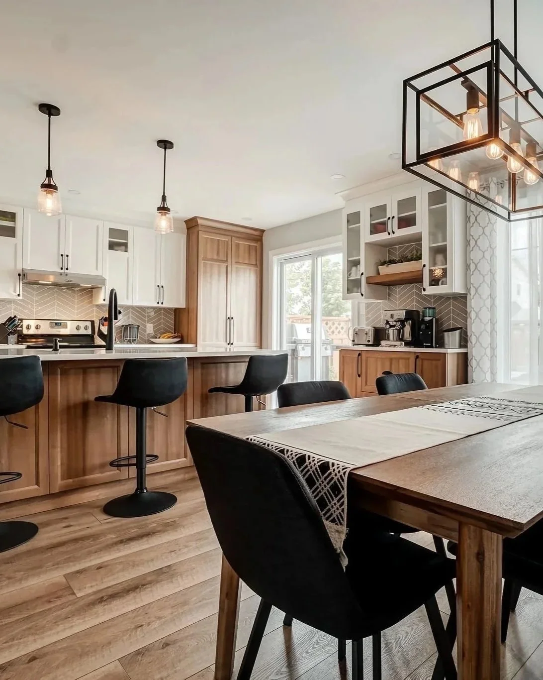 Cuisine moderne avec bar en bois, chaises noires, armoires blanches, étagères en bois, appareils électroménagers, luminaires suspendus et luminaires encastrés près d'une porte-fenêtre.