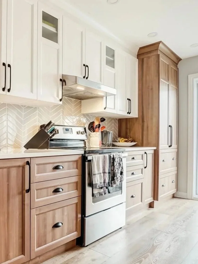 Cuisine avec armoires en bois clair et blanc, évier, four électrique, dosseret en carreaux beige et blanc à motif de chevrons, plan de travail en quartz, ustensiles de cuisine, torchons suspendus sur le four.