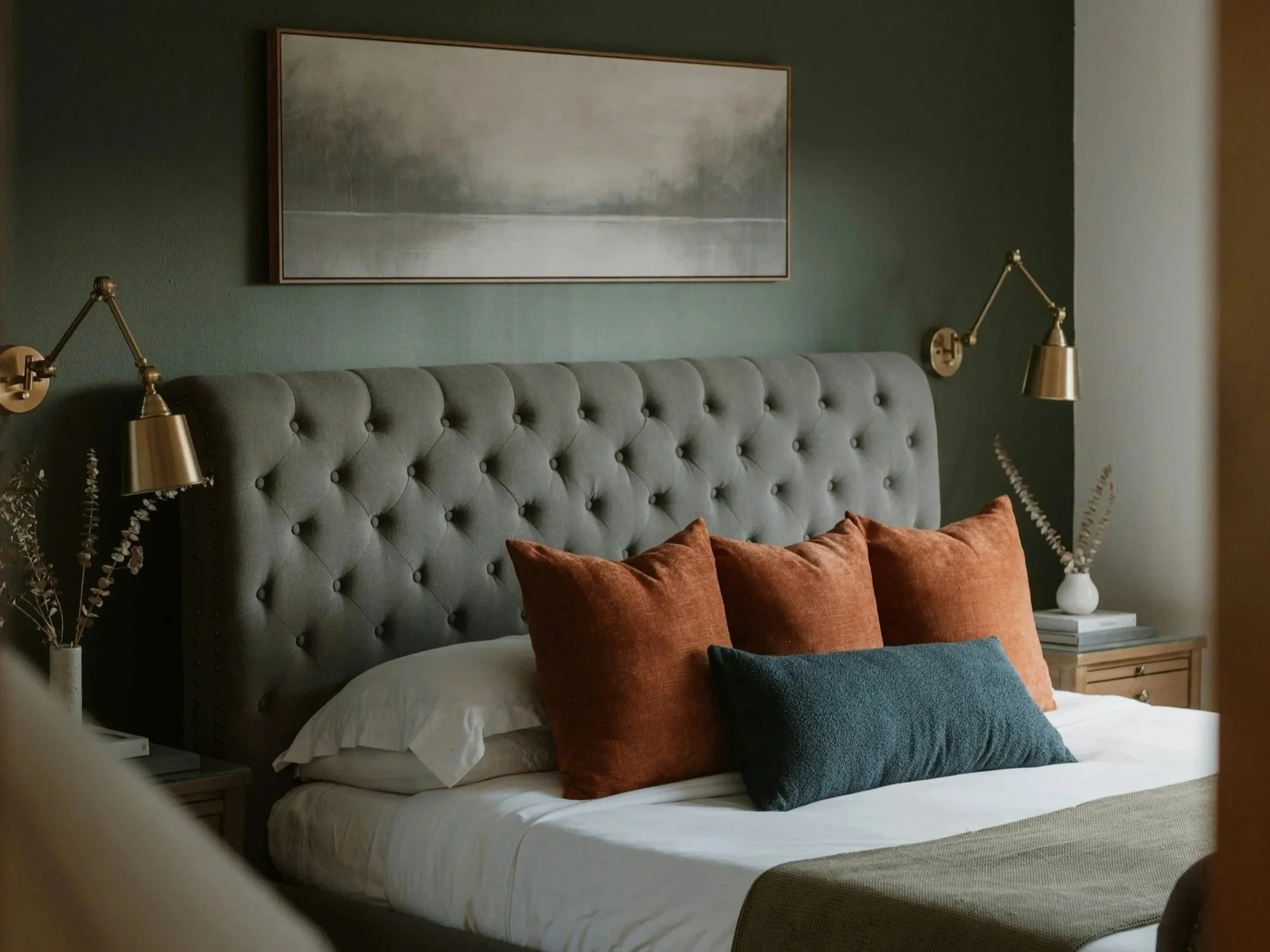 Chambre à coucher avec un lit à tête capitonnée grise, des coussins orange et un coussin bleu, deux lampes de lecture dorées, une toile abstraite au-dessus du lit, et deux tables de chevet avec des vases de fleurs séchées.