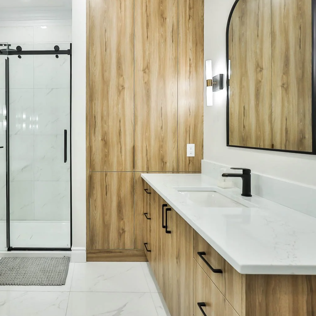 Une salle de bain moderne avec un meuble en bois clair, un miroir rectangulaire avec cadre noir, un évier intégré en marbre blanc, un robinet noir, un mur avec un luminaire mural et un mur en bois, une porte de douche en verre avec cadre noir et une