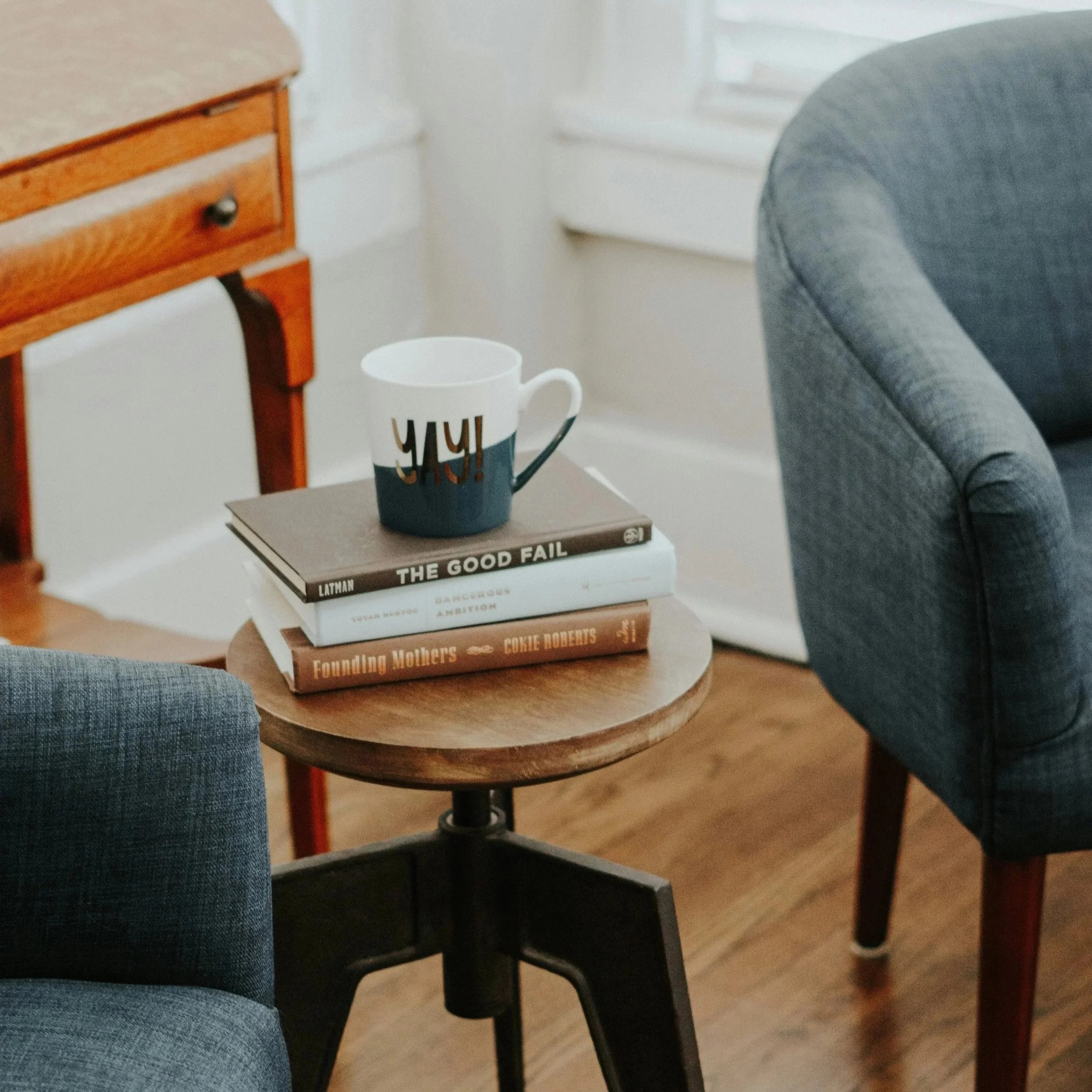Une tasse posée sur un livre sur une petite table en bois, entourée de deux fauteuils en tissu gris, dans une pièce lumineuse.