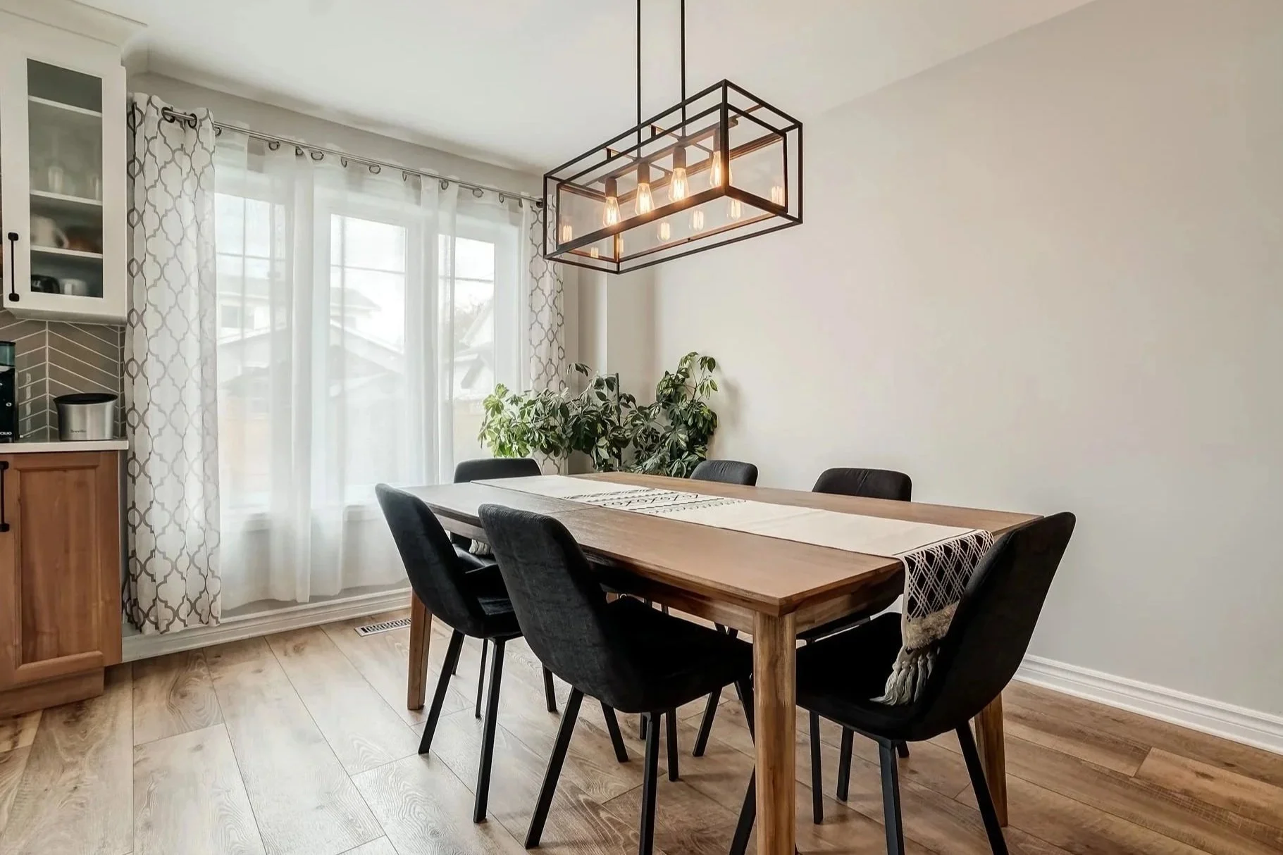 Salle à manger avec une table en bois, six chaises noires, rideaux blancs à motifs gris, un luminaire suspendu avec plusieurs ampoules et des plantes en arrière-plan.