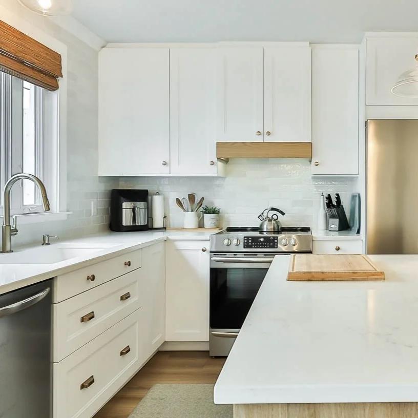 Cuisine campagne moderne lumineuse avec comptoirs en pierre blanche, armoires blanches, un évier, un four , un réfrigérateur en acier inoxydable, une fenêtre avec stores en bois, et plusieurs ustensiles de cuisine.