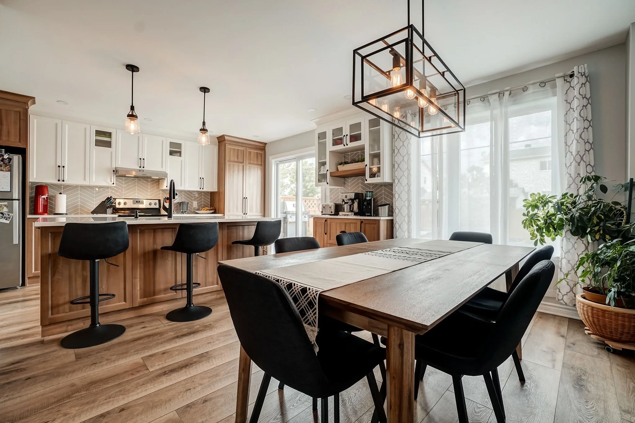 Cuisine moderne contemporaine avec plan de travail en bois, chaises noires, luminaire industriel au plafond, grande baie vitrée avec rideaux blancs, plantes d'intérieur.