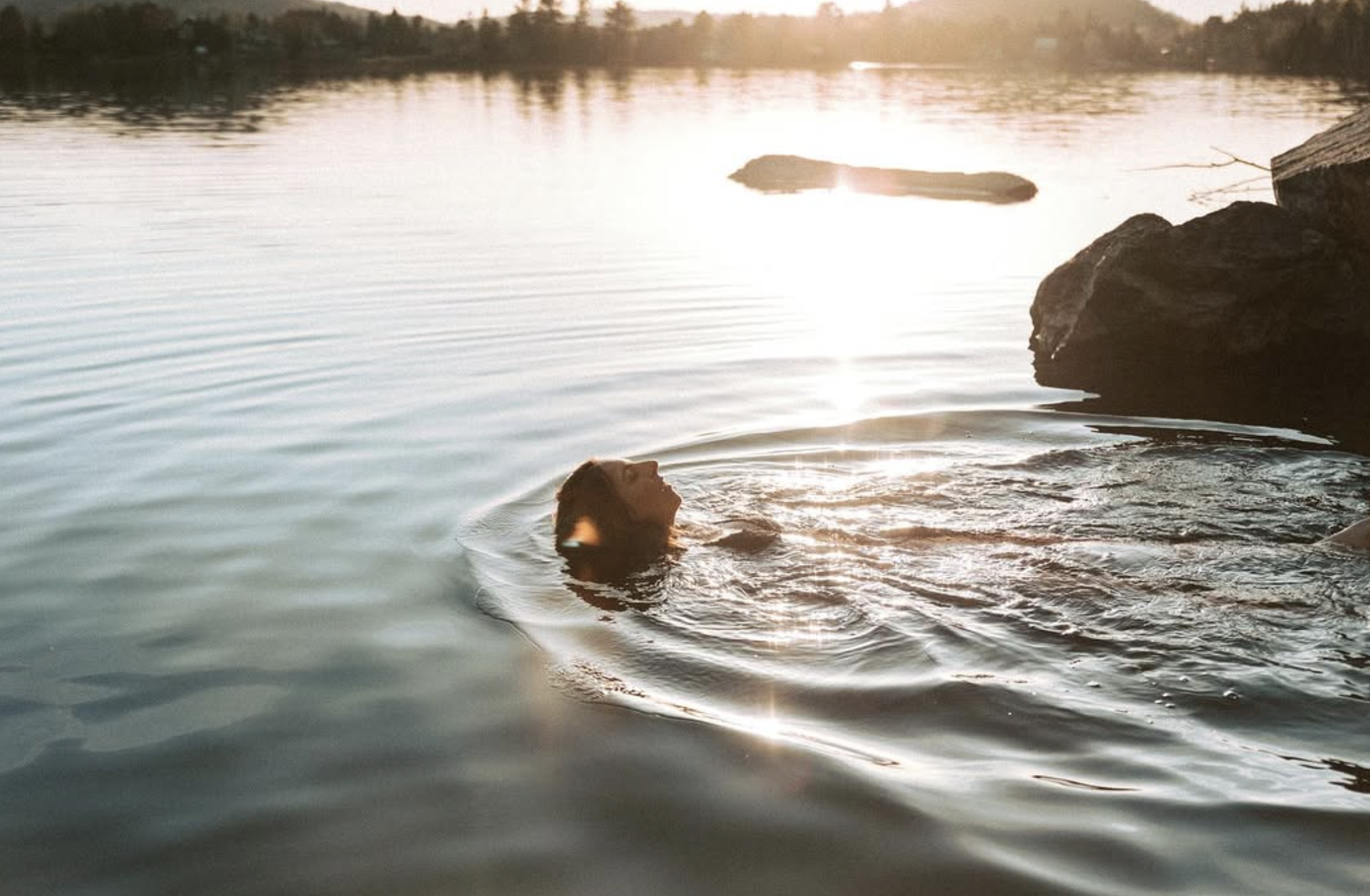 Une femme assise à la fin d'un quai en bois, regardant l'eau, portant un bonnet gris, une veste grise et des chaussures jaunes et noires, avec ses cheveux longs et bruns.