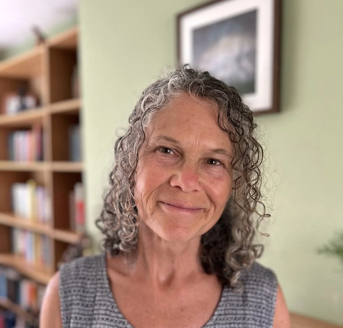 Another photo of Mandy Marty, the Cambridge-based counsellor, with curly hair smiling, standing in a room with a bookshelf and a framed picture on a light green wall.