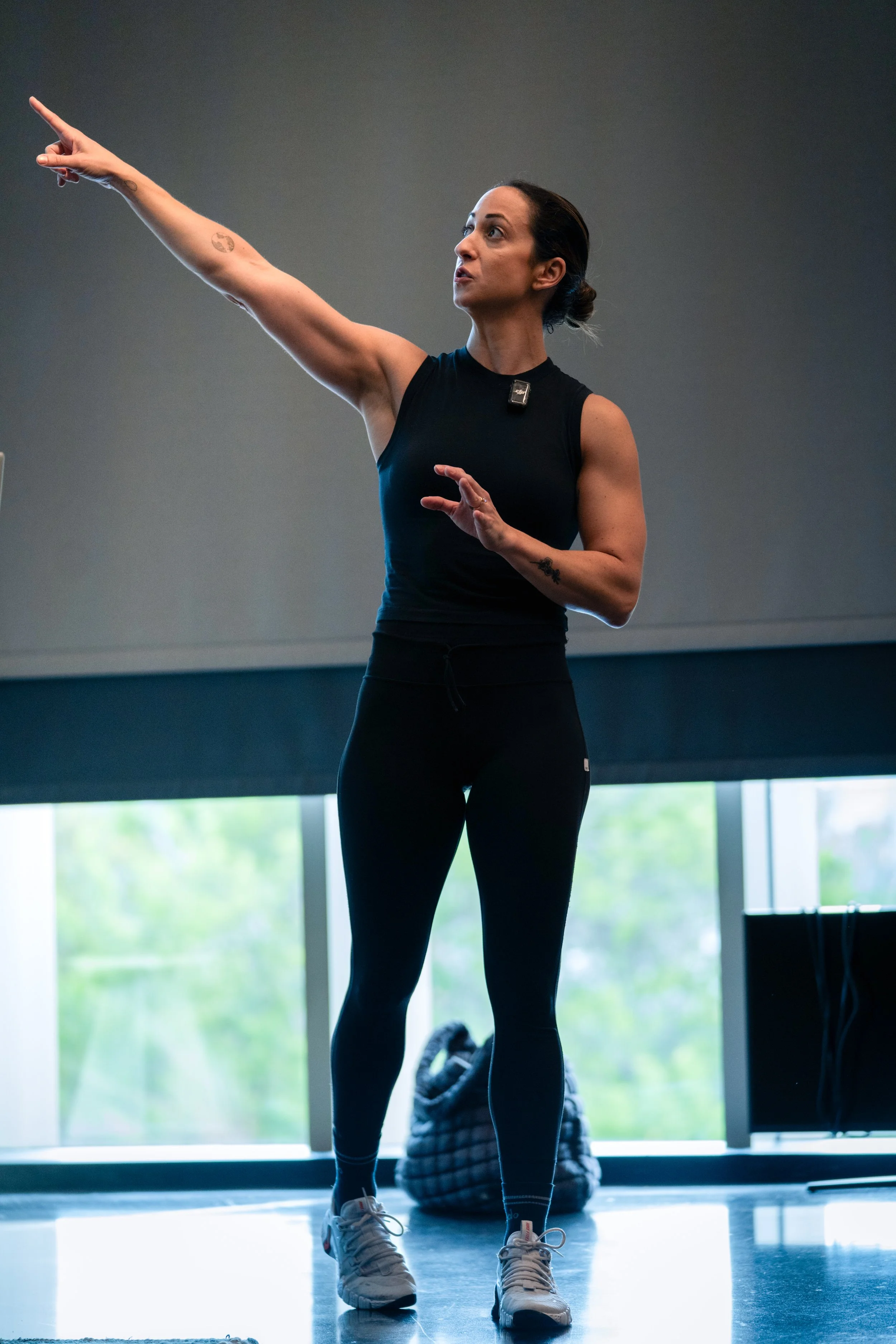 Woman in black sleeveless top and leggings giving a presentation or instruction in a gym or fitness studio, pointing to the left with her right arm, while standing in front of large windows with greenery outside.
