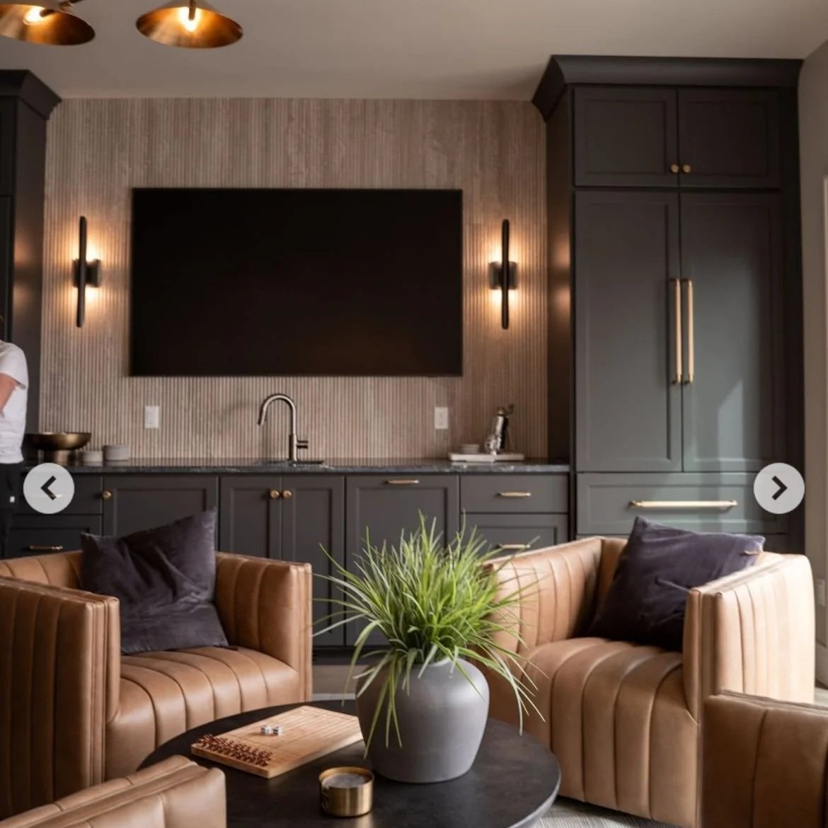 Living room with beige leather armchairs, dark cabinetry, a large TV, and modern wall sconces.