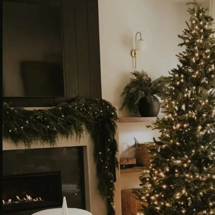 A decorated Christmas tree with lights in a living room with a fireplace, a green garland on the mantle, and a black vase with greenery on a shelf.