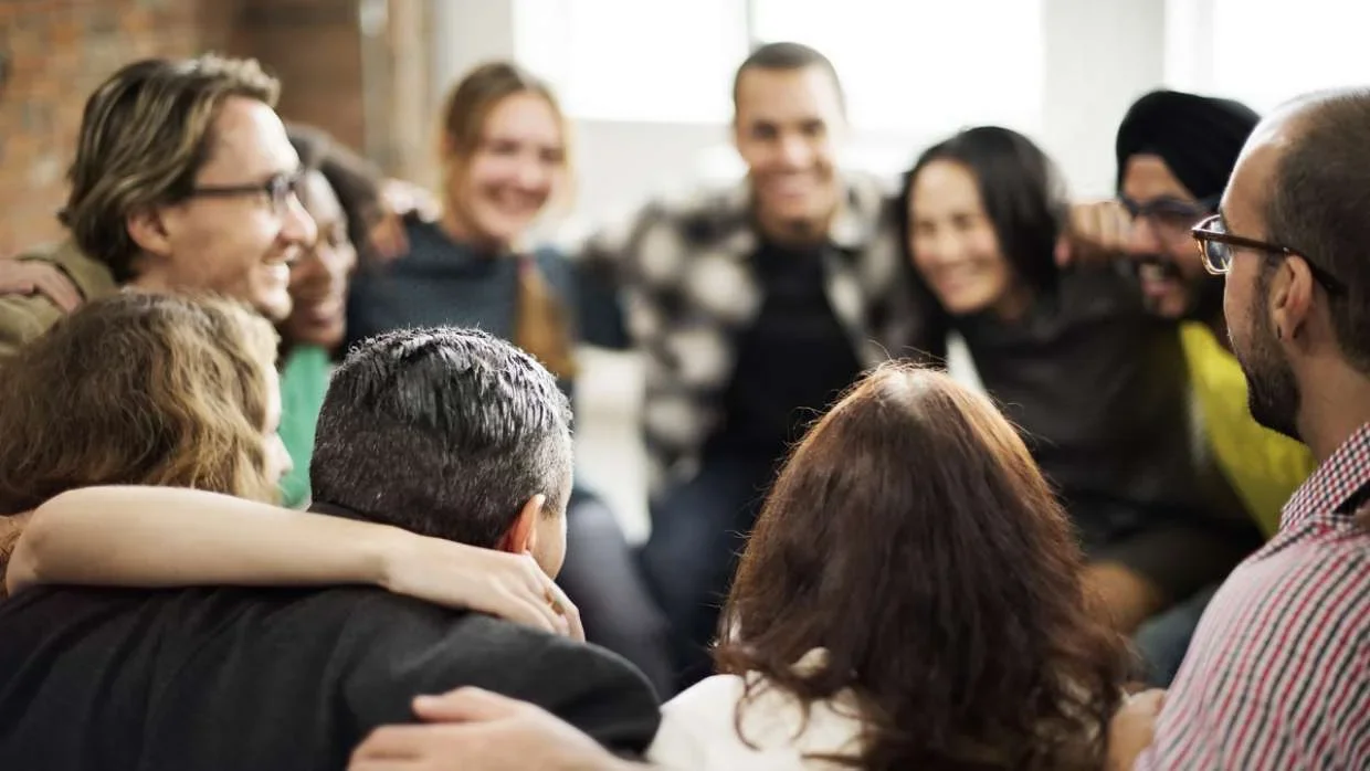 A diverse group of people sitting closely together in a circle, smiling, and hugging, in a bright room with natural light.