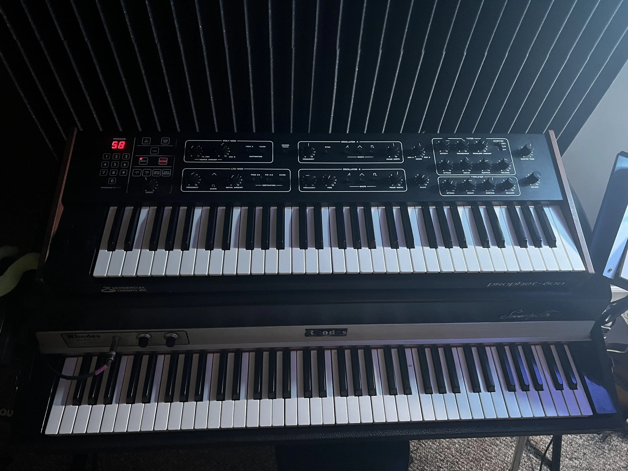 A musical setup featuring an upper synthesizer keyboard and a lower piano, positioned on a stand against a dark background with vertical lines.