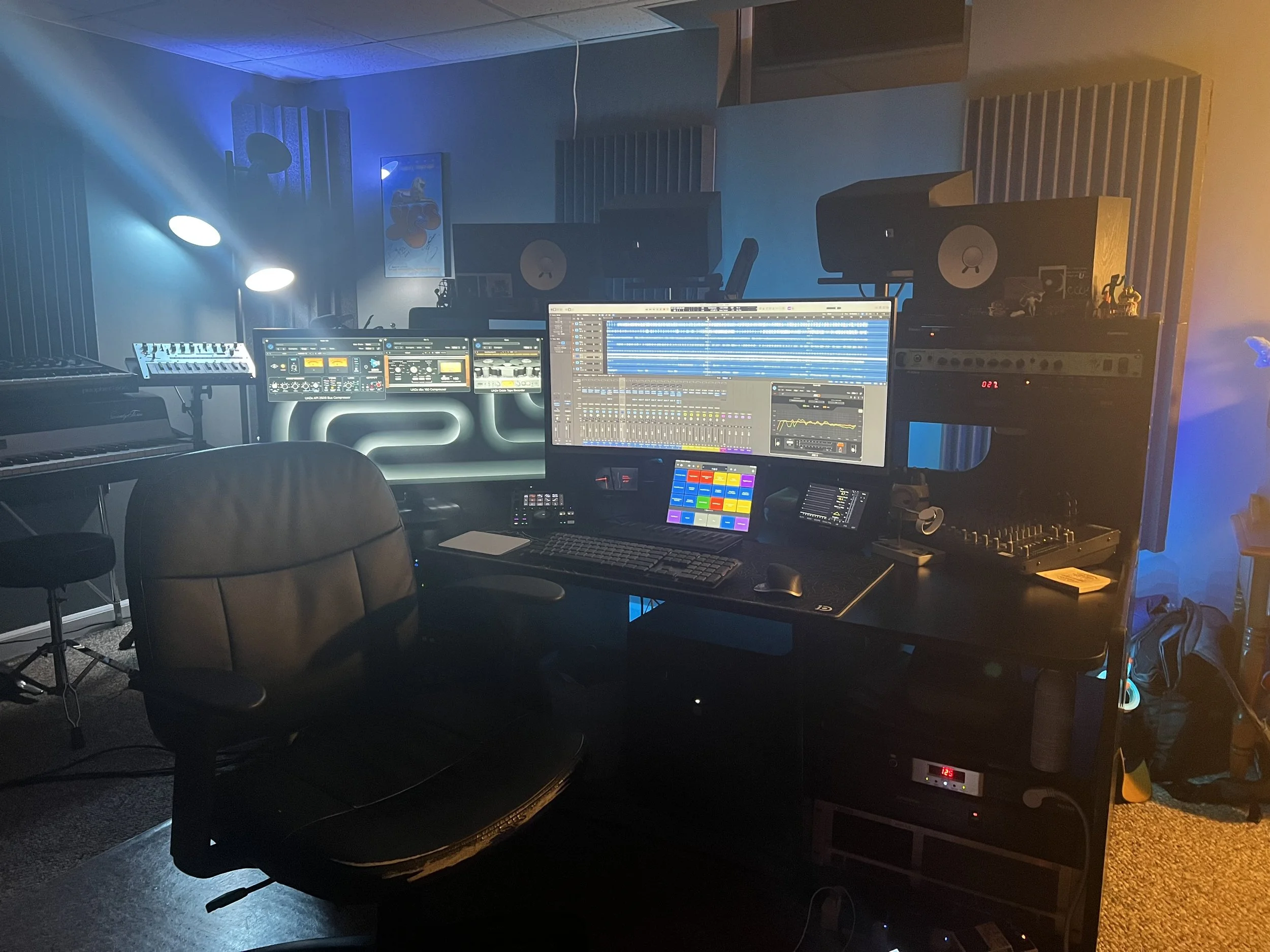 A music production studio with multiple computer screens, audio equipment, and studio monitors, illuminated with blue and purple lighting.