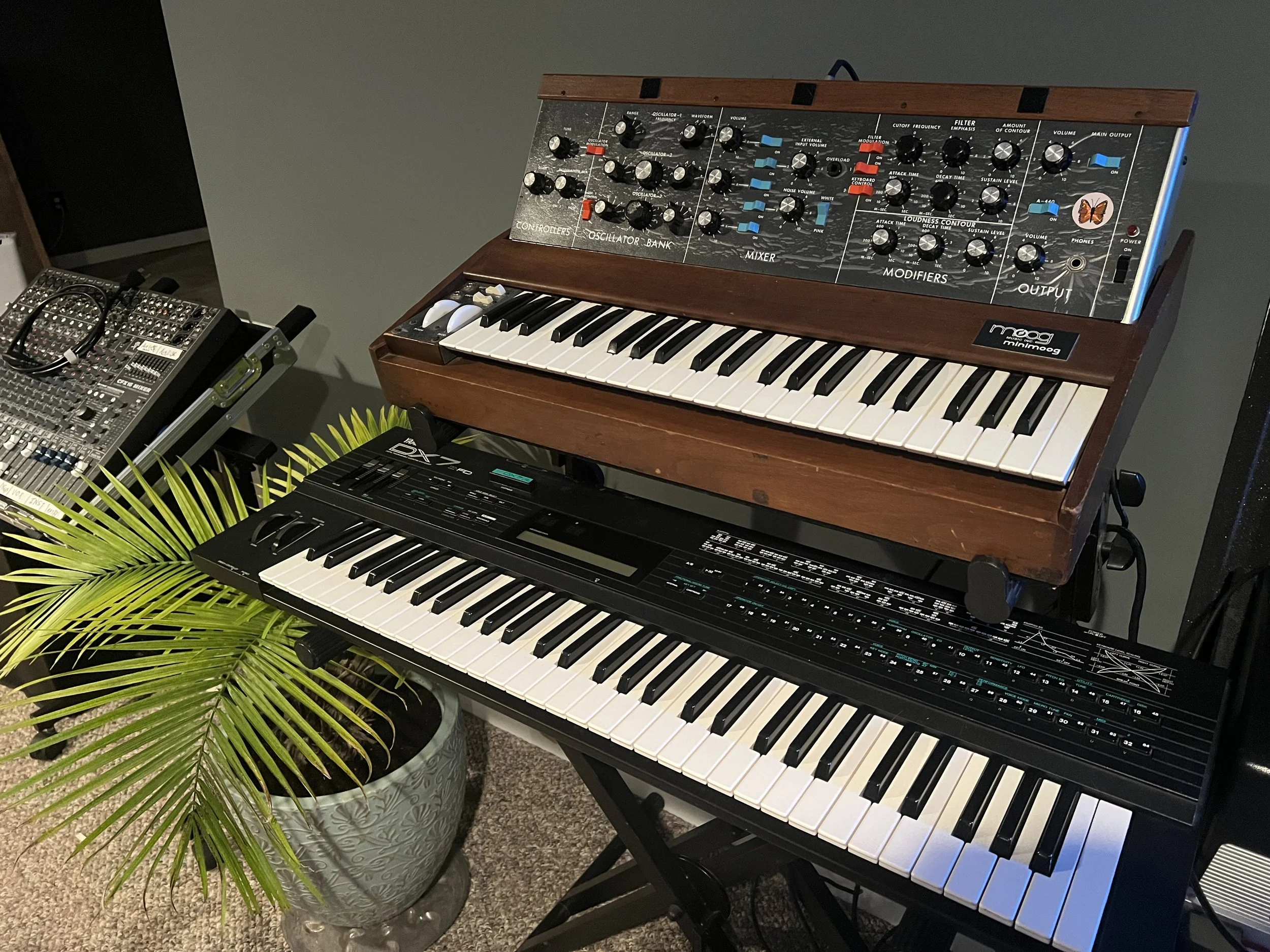 Two electronic and one analog keyboard synthesizers, a mixing console, and a potted plant in a room with gray walls.