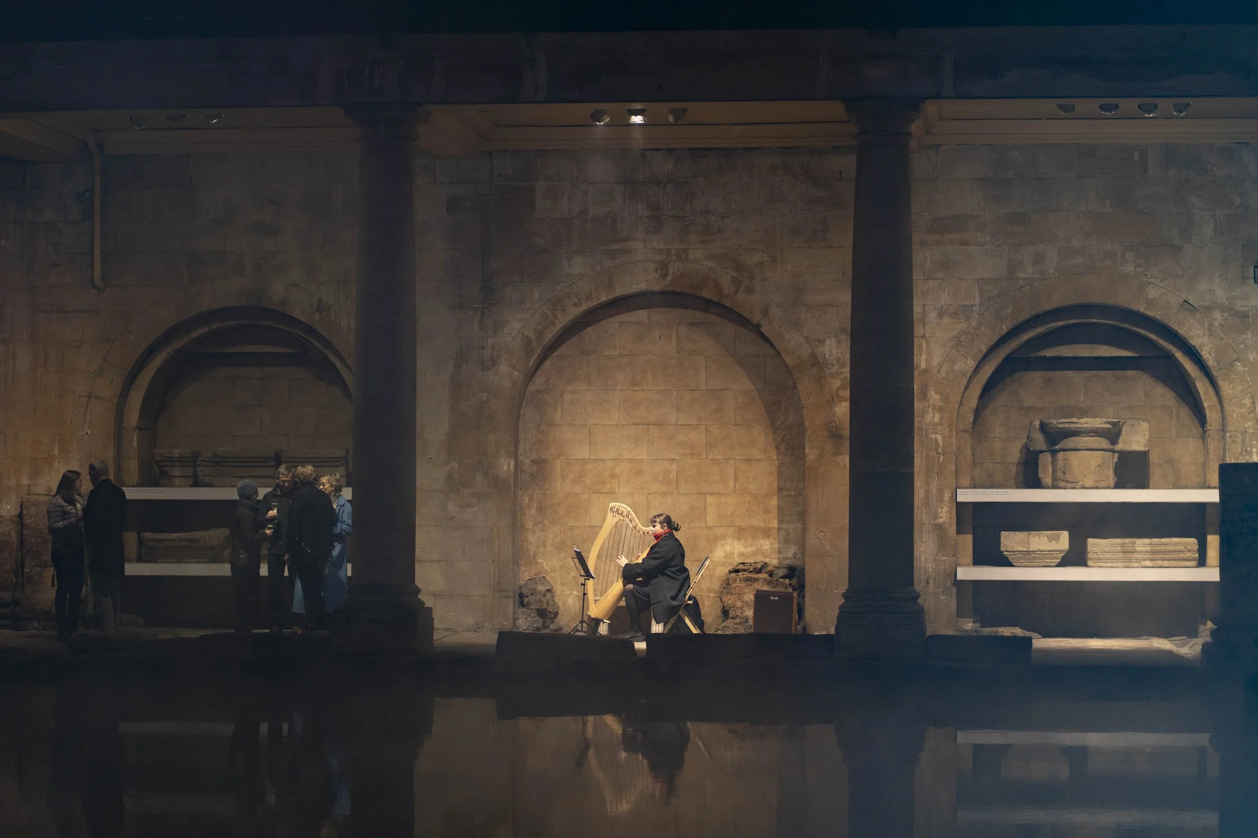 A harpist performing on a small stage in a historic building with stone walls and arches, while a group of people observe nearby.