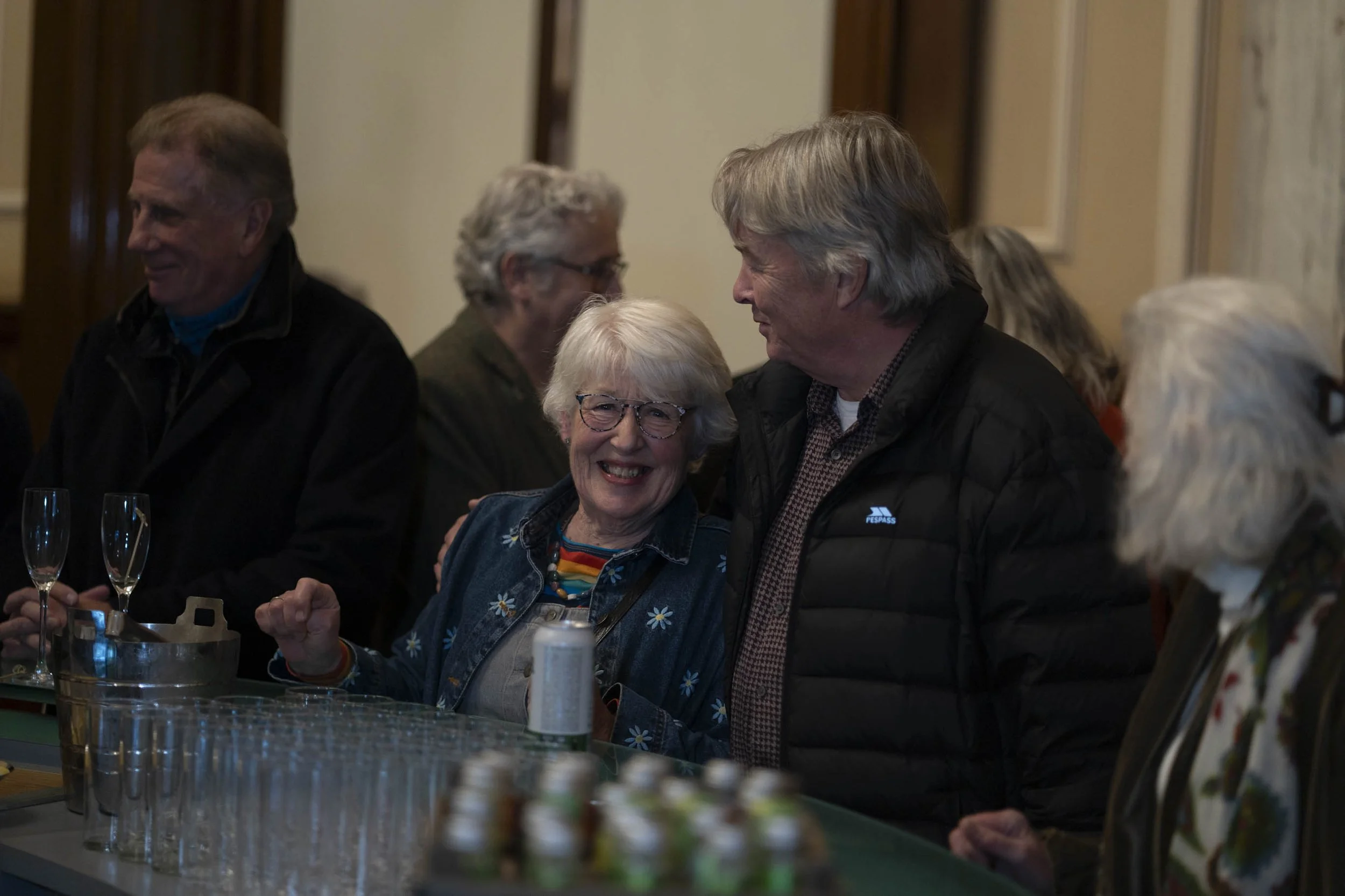 A group of smiling older adults at a social gathering, mostly gathered around a bar or table with drinks, engaging in conversation, with one woman and man in the center sharing a happy moment.