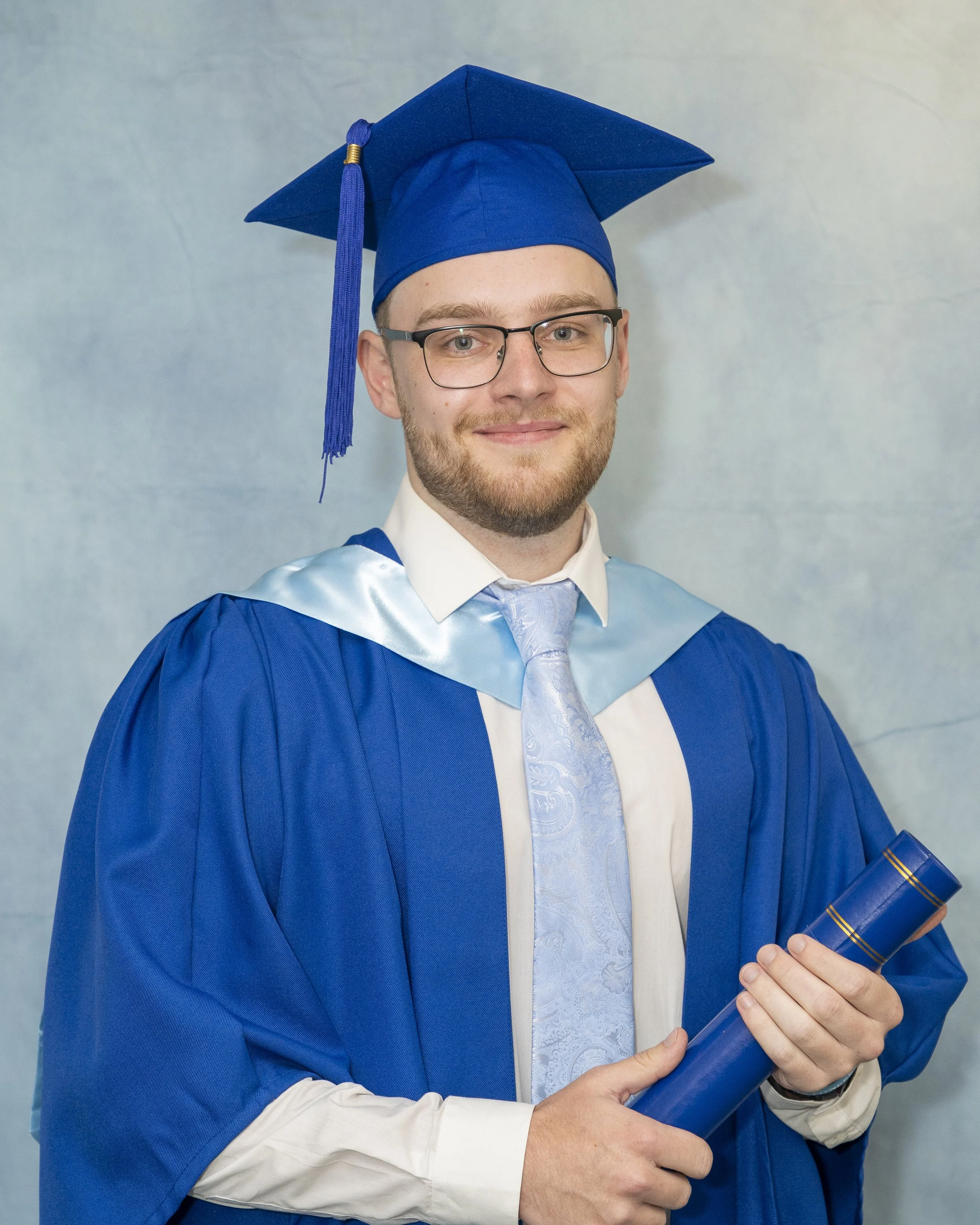 A young man in a blue graduation gown and cap, holding a diploma, smiling at the camera.