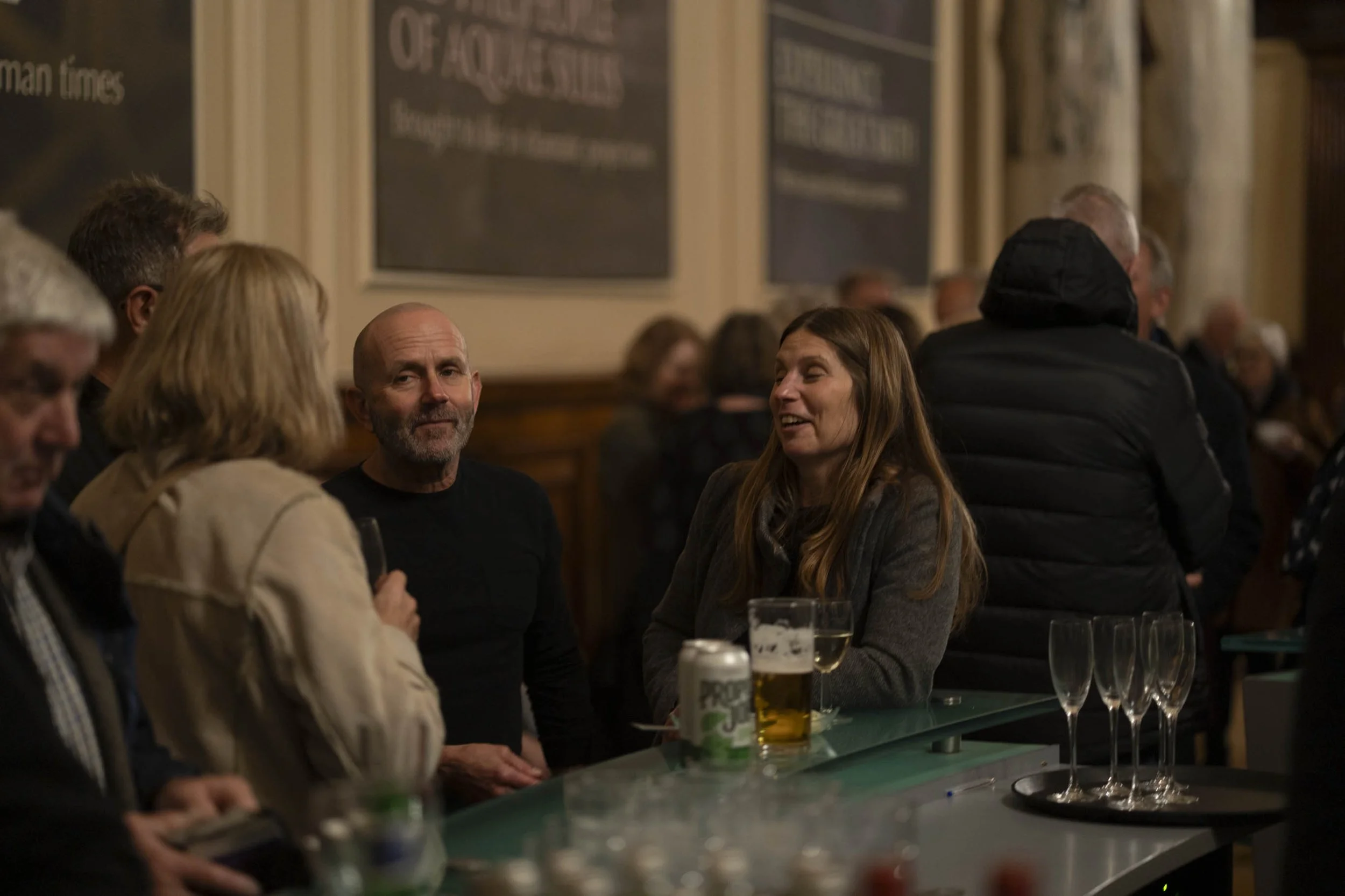 People socialising at a bar or pub, with drinks on the counter, in a dimly lit indoor setting.