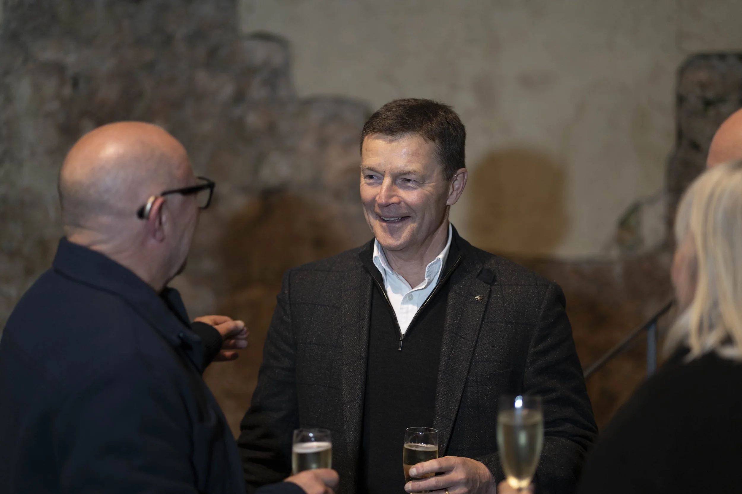 Three people at a social gathering, two men and one woman, are having a conversation, holding glasses of champagne, in an indoor setting with a masonry wall background.