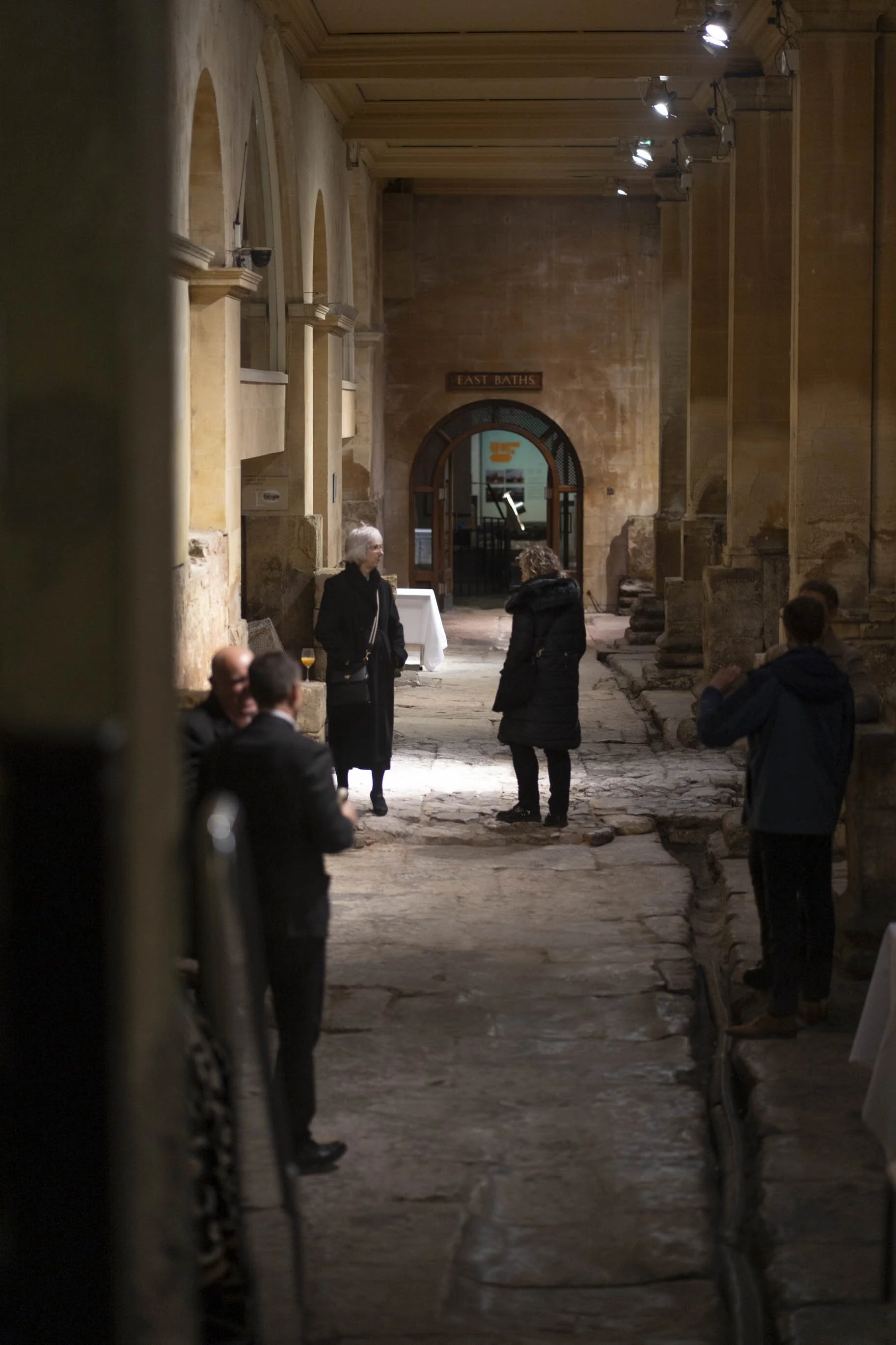 People gathered in Roman Baths corridor with a sign that reads 'Fast Baths', some engaged in conversation, and a table with drinks visible in the background.