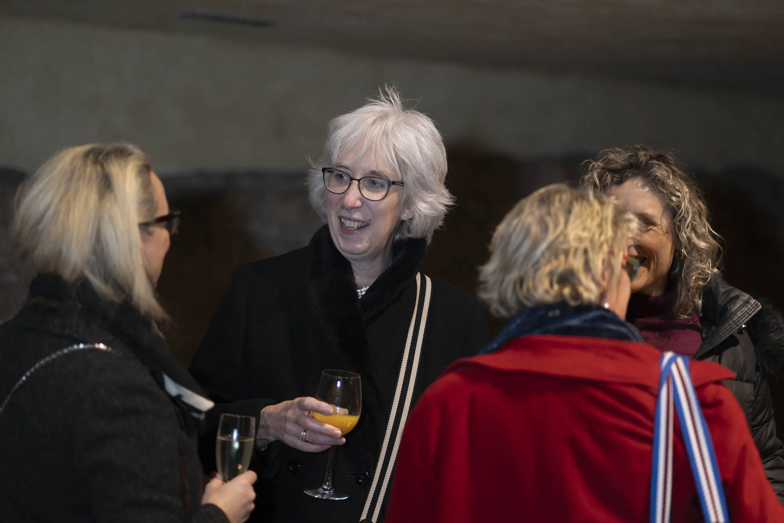 Group of four women talking at a social gathering, some holding drinks, with one woman wearing glasses and a black coat, smiling.