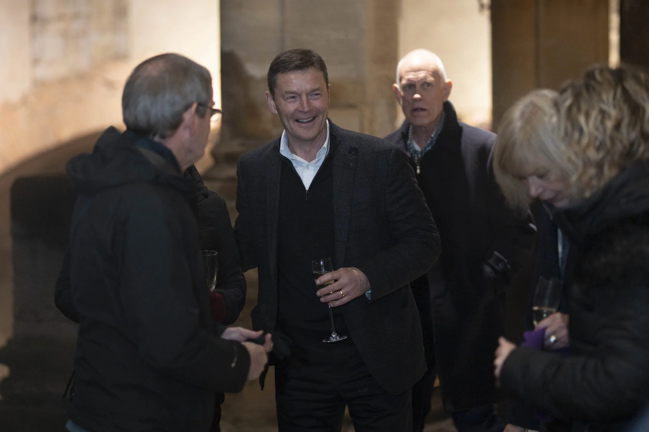 Group of people socialising indoors, with one man in the center holding a champagne flute, smiling and engaging in conversation at Roman Baths.