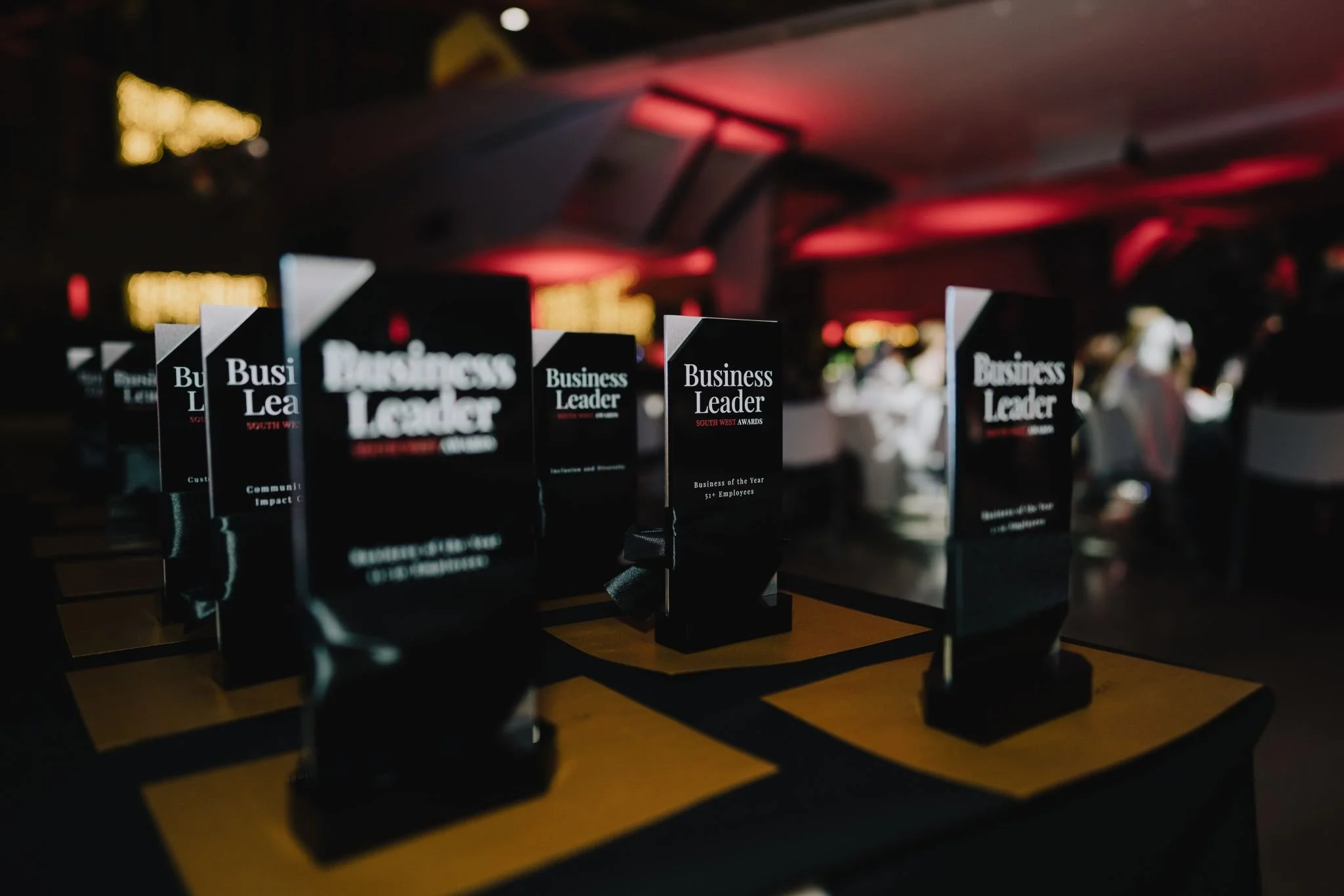 Business awards plaques on a table at an event with blurred background of people and red lighting.