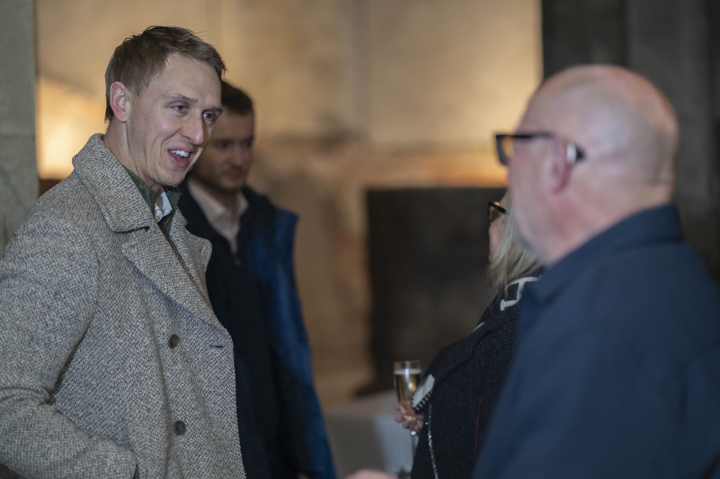 Four people engaged in conversation at a social gathering. One man in a beige coat talking to a woman holding a glass of champagne, while two men stand in the background at Roman Baths.