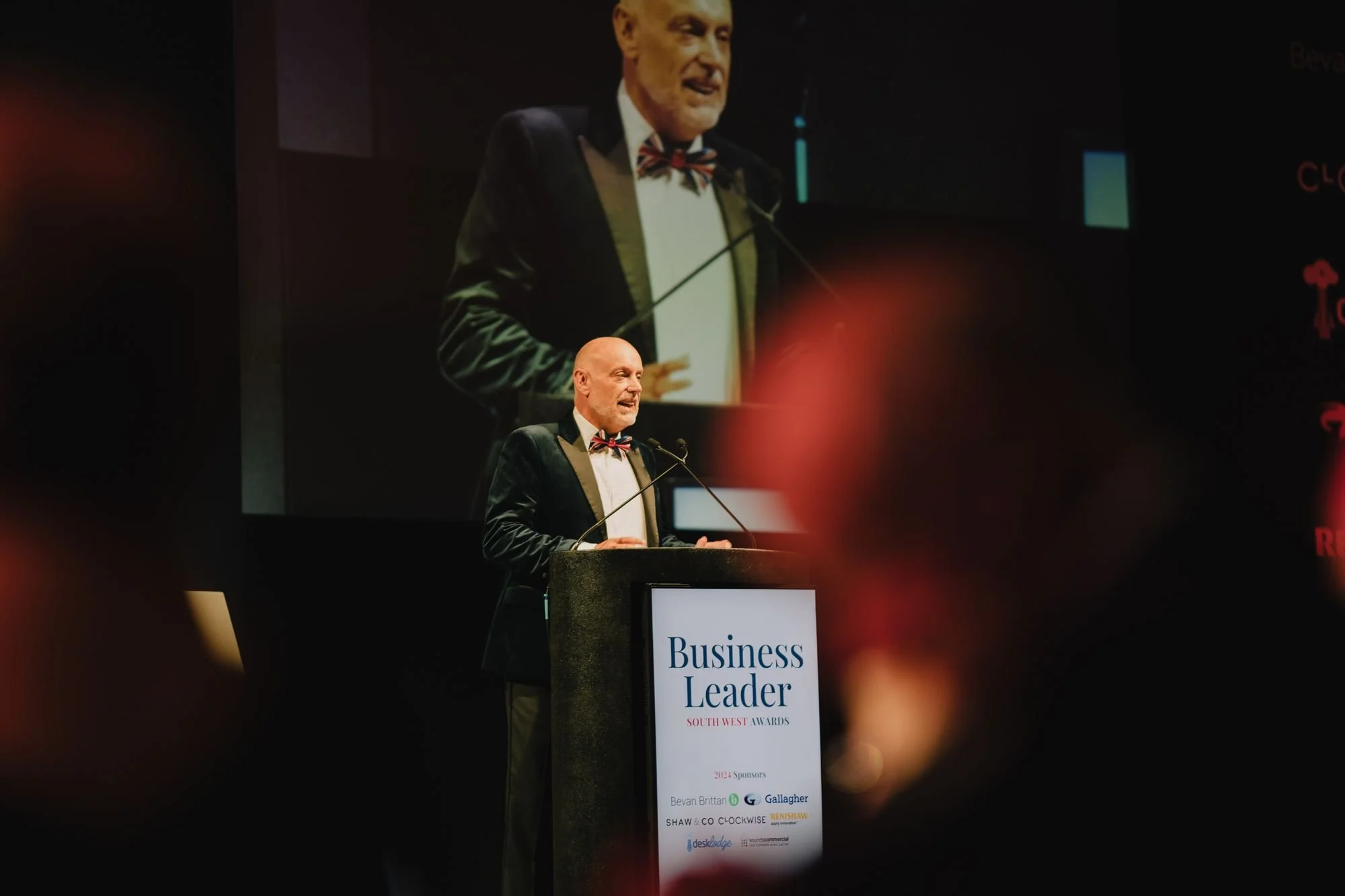 Man in tuxedo with bow tie speaking at a podium during a business awards event, with a large screen behind him displaying his image.
