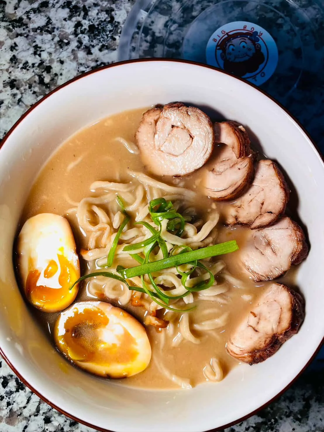 A bowl of ramen noodles with sliced pork, soft-boiled eggs, green onions, and broth on a speckled countertop.