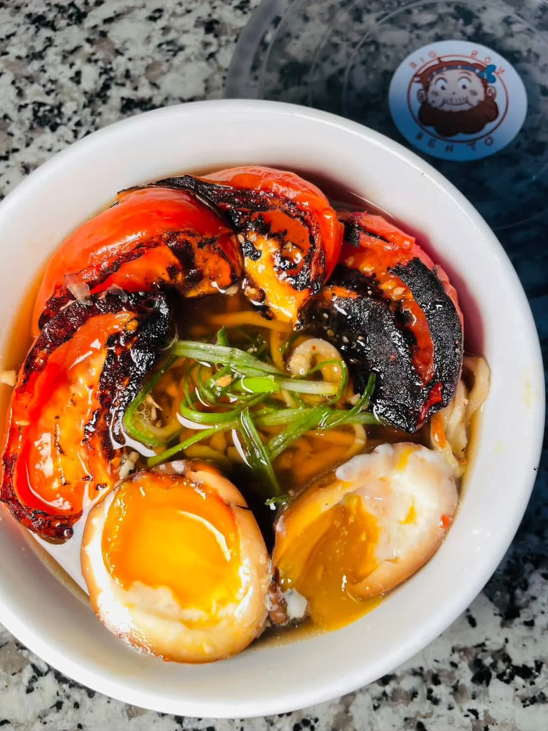 A bowl of ramen noodles with grilled tomatoes, soft-boiled eggs, and green onions.