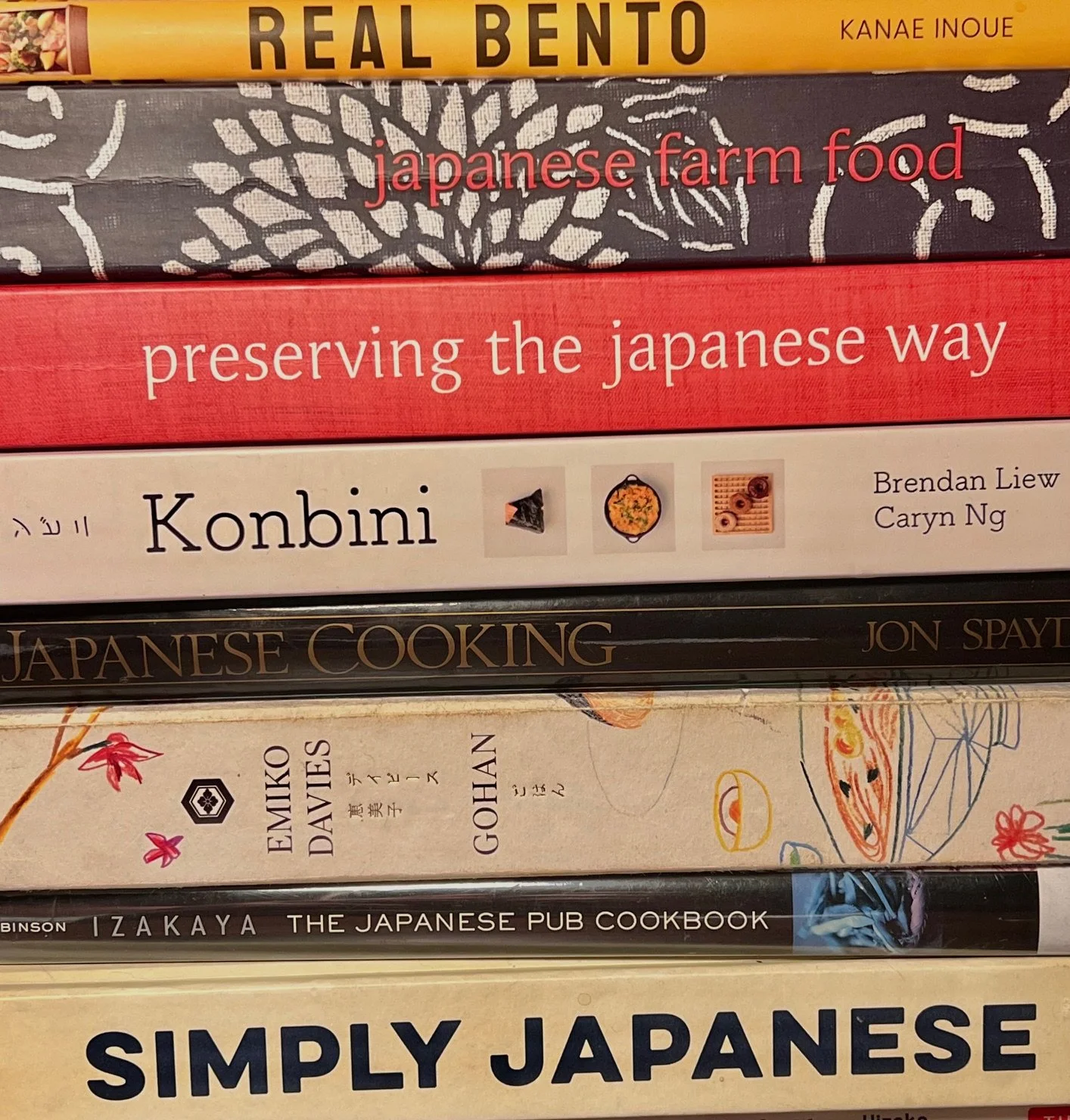 Stack of books about Japanese cuisine and culture, including titles like 'Real Bento,' 'preserving the Japanese way,' 'Konbini,' 'Japanese cooking,' 'Emiko Davies,' 'Izakaya,' and 'Simply Japanese.'